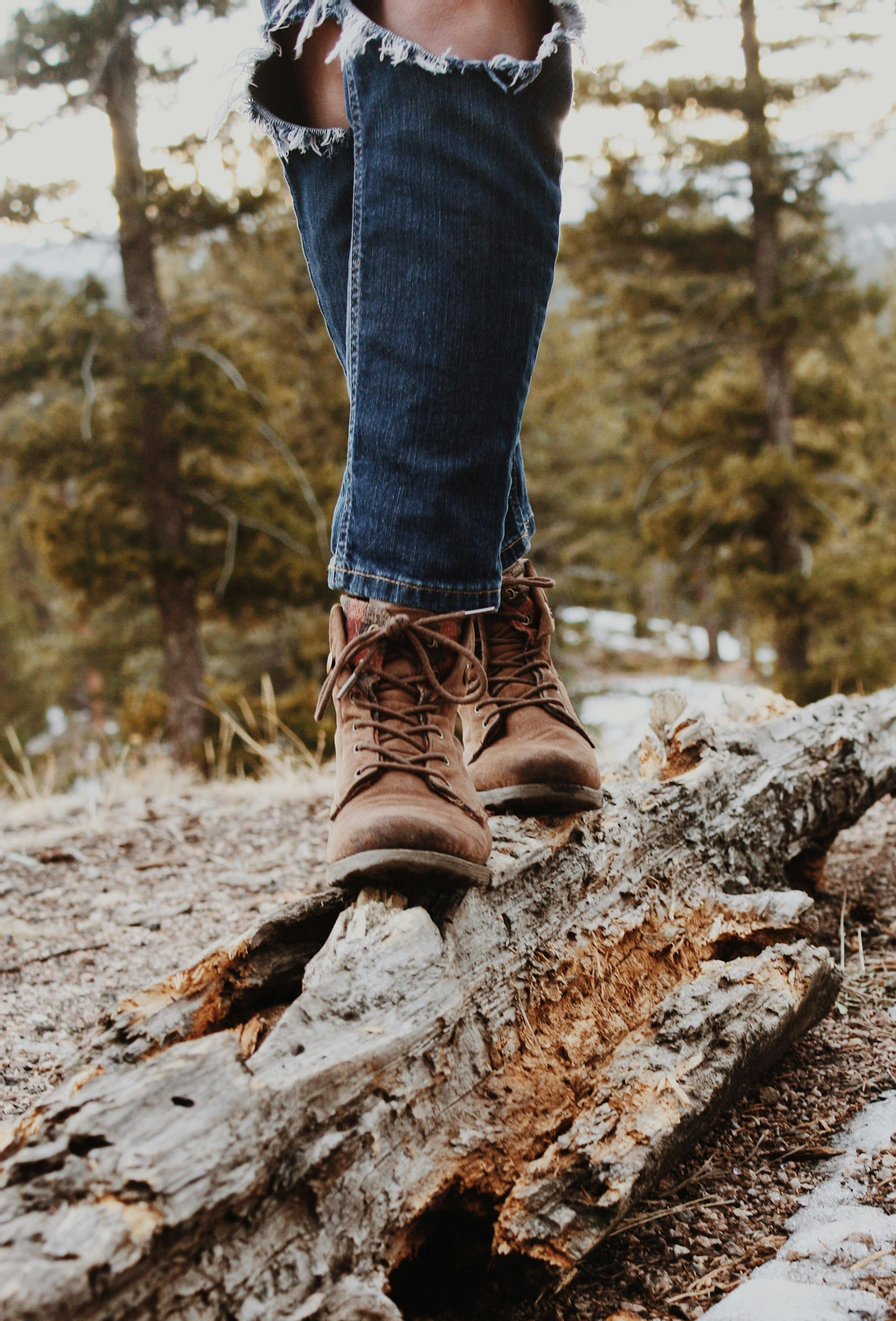 Person wearing ripped jeans and brown boots balancing on a fallen log in a forested area.