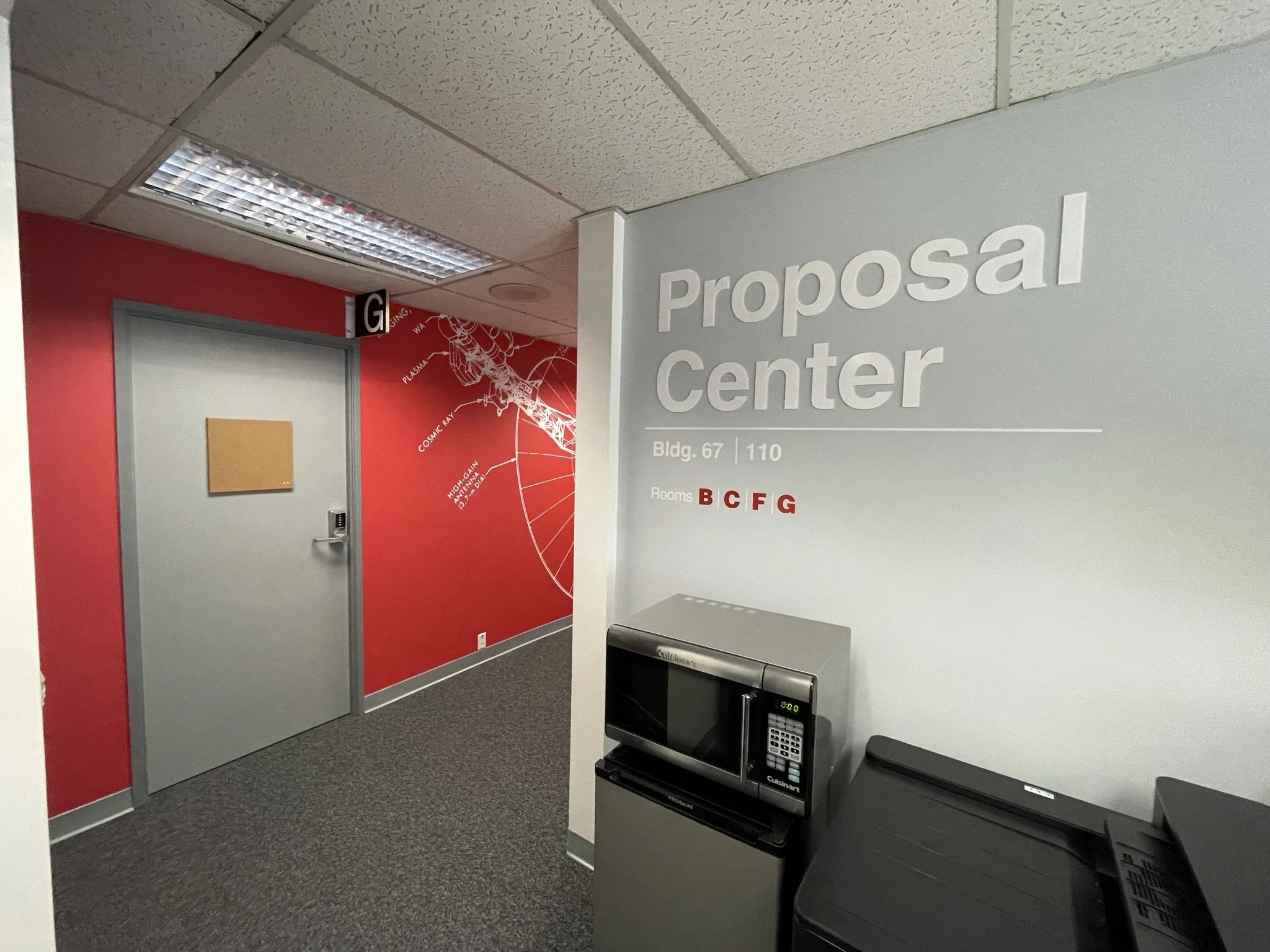 Office hallway with red and gray walls, a door on the left, a large 'Proposal Center' sign on the right, and a microwave and office phones on a counter in the foreground.