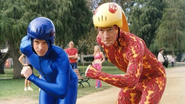 Two men in colorful costumes posing dramatically outdoors, with a park and onlookers in the background.