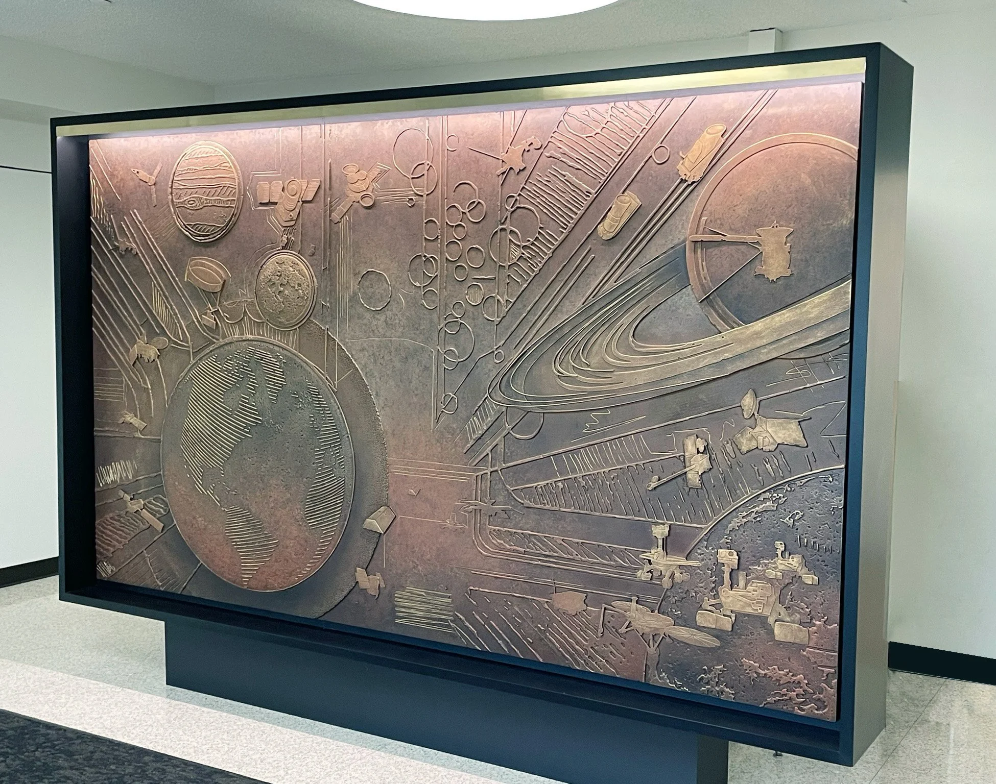 Framed artwork depicting a stylized map of the world, scientific instruments, and abstract lines and circles, displayed against a wall in a gallery.