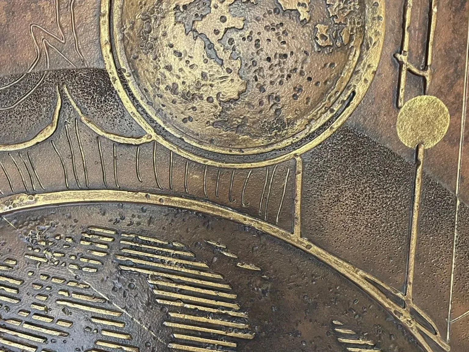 Close-up of a textured metallic artwork featuring circular shapes, lines, and a compass-like motif with a gold and dark bronze color palette.