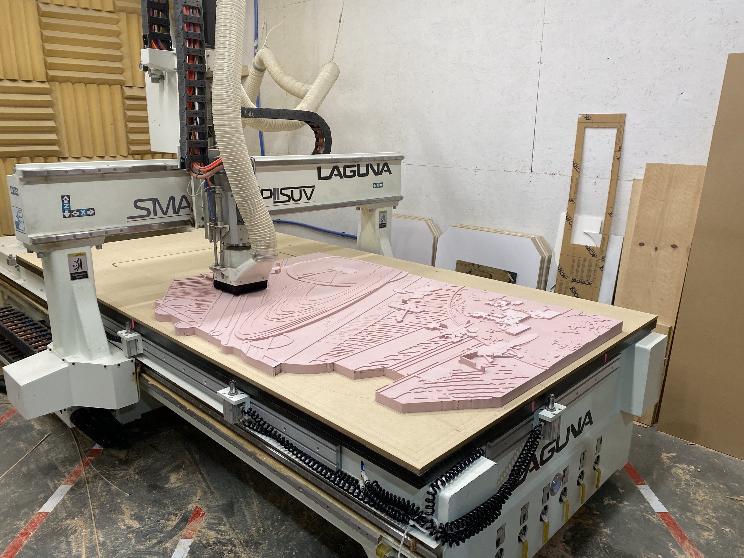 A CNC machine working on a pink foam board with a detailed surface map or architectural model, in a workshop with foam pieces and tools around.