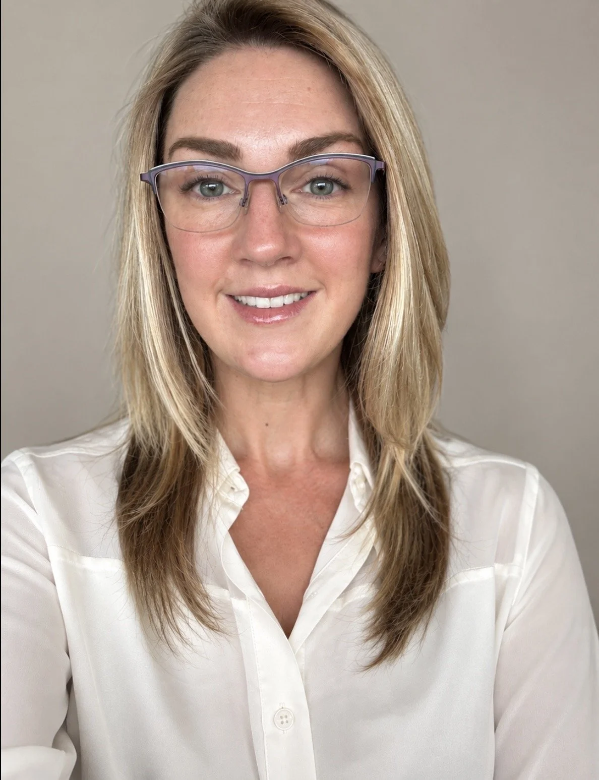 A woman with shoulder-length blonde hair, wearing glasses and a white button-up shirt, smiling at the camera against a plain beige background.