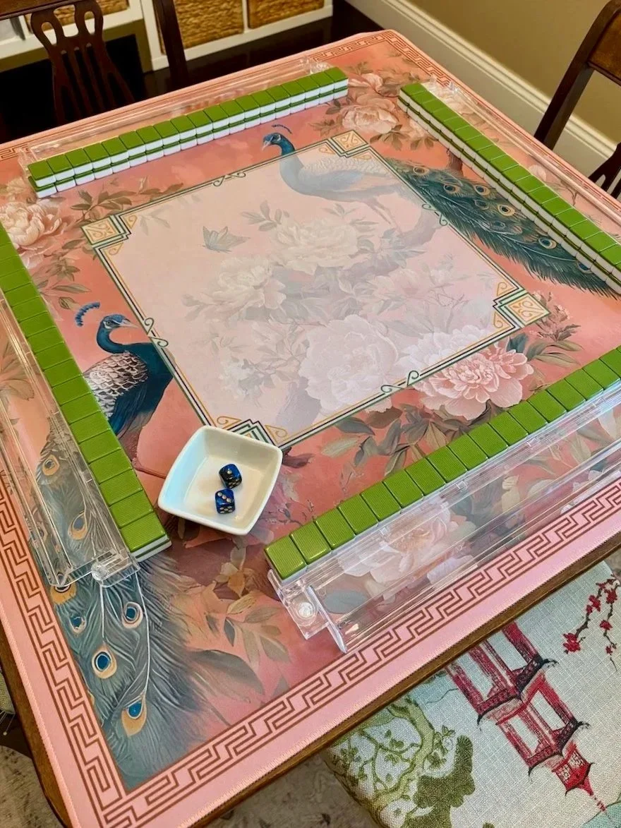 A Mahjong table with a floral design and peacock images on the surface, green tiles arranged around the edges, and a white dish with two blue dice on it.