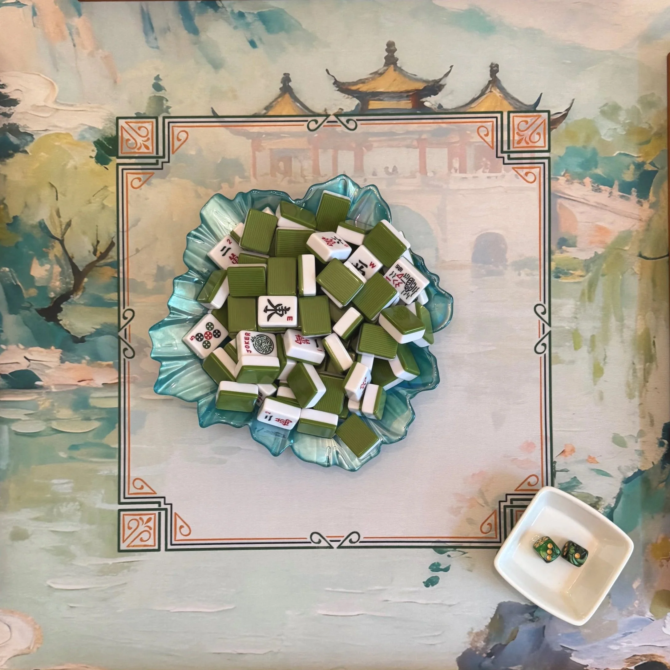 A table with a decorative border features a blue glass dish filled with Mahjong tiles and a small white dish with two dice.