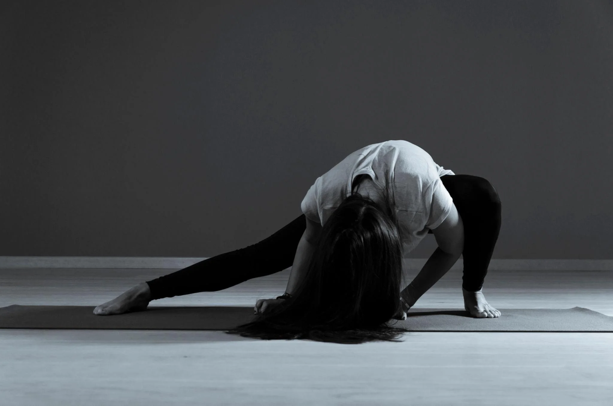A person with long hair practicing yoga indoors, stretching on a mat in a lunge position with one leg extended and the other bent, against a plain dark wall.