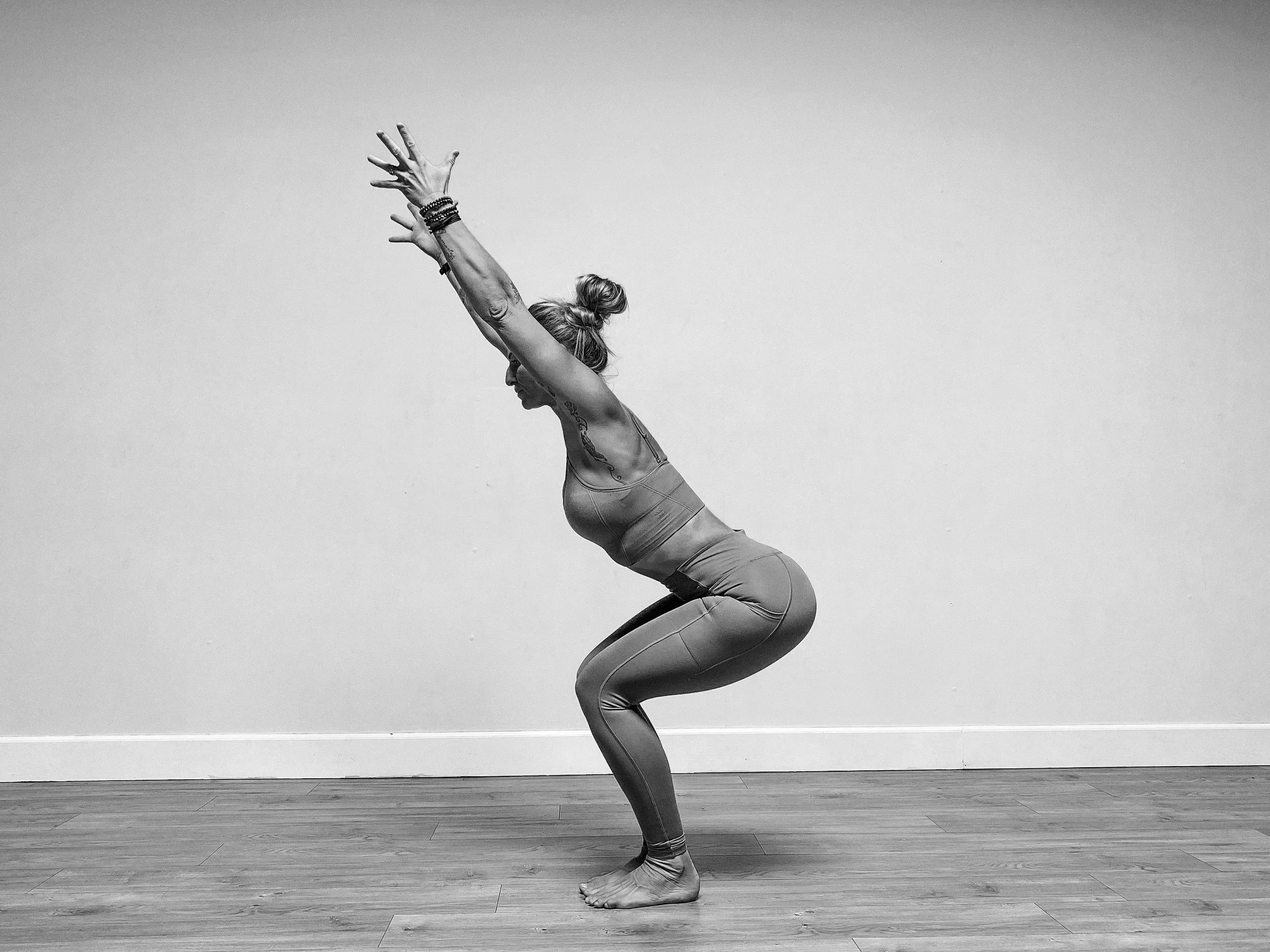 A woman practicing yoga indoors, captured in a side profile, in a squat position with her arms raised above her head, against a plain wall and wooden floor.
