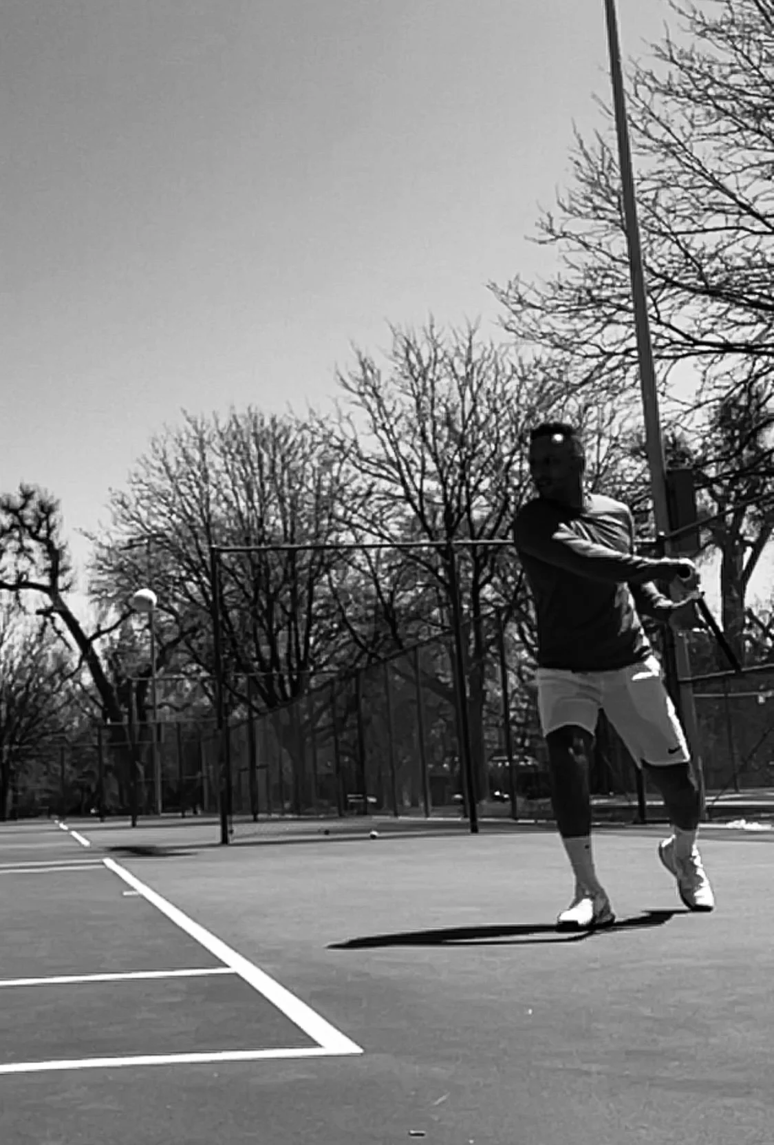 A person playing tennis on an outdoor court with trees and a fence in the background, black and white photo.