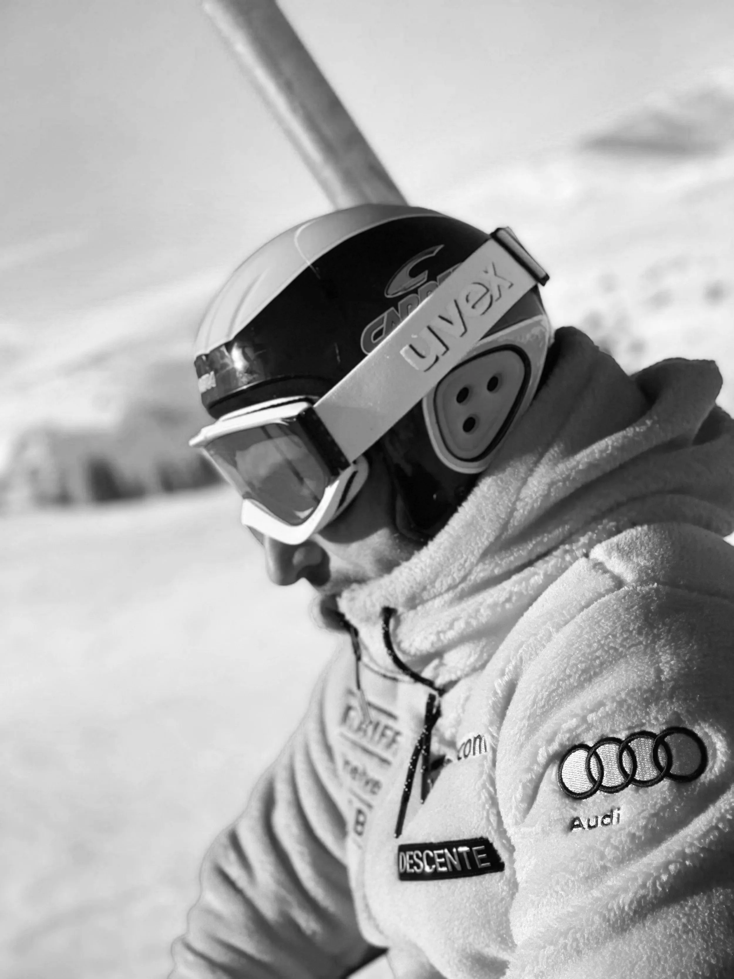 A skier wearing a helmet and goggles, dressed in a jacket with patches, standing outdoors in a snowy landscape.