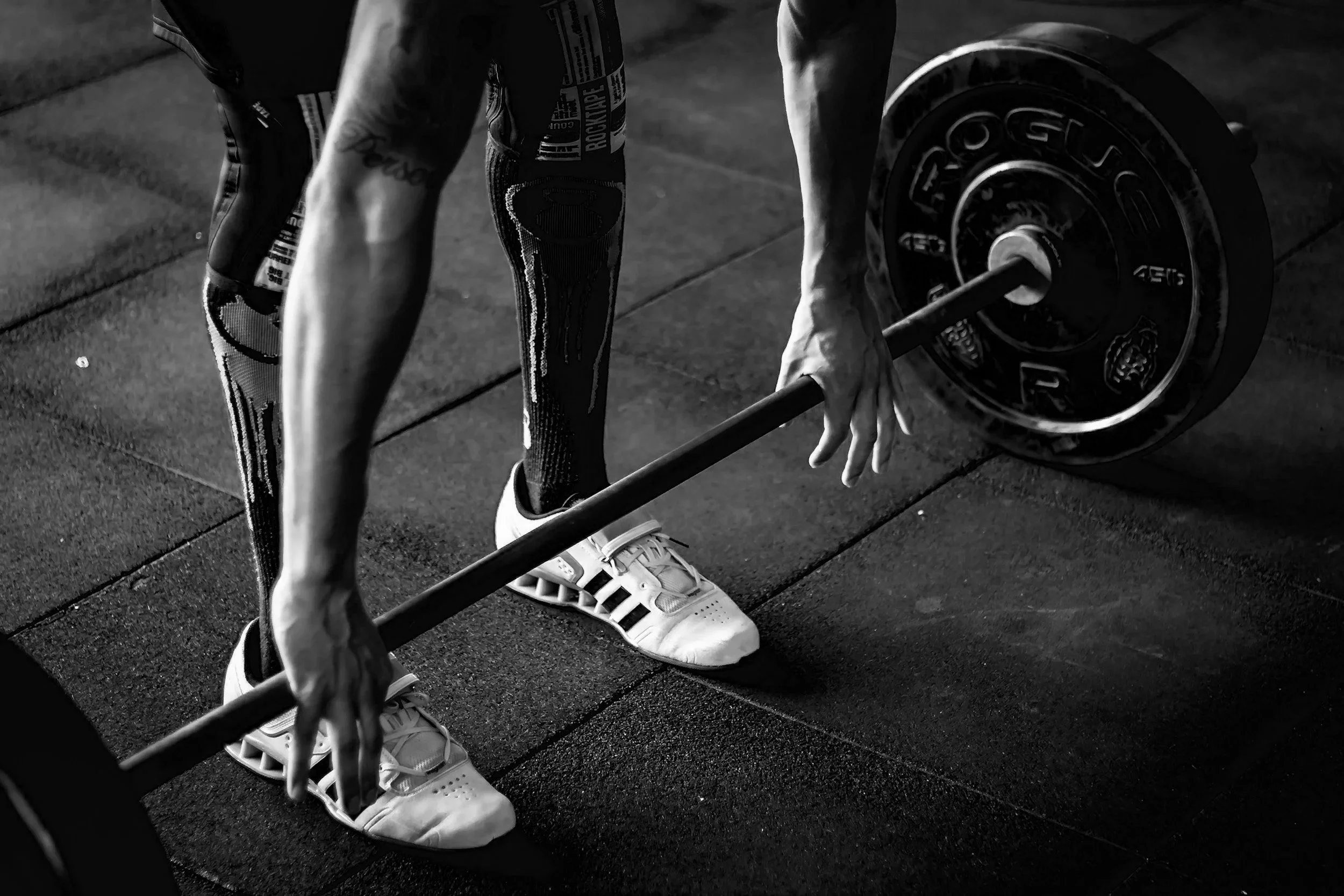 A person gripping a barbell with weight plates, wearing athletic shoes and knee-high compression socks, on a gym floor.