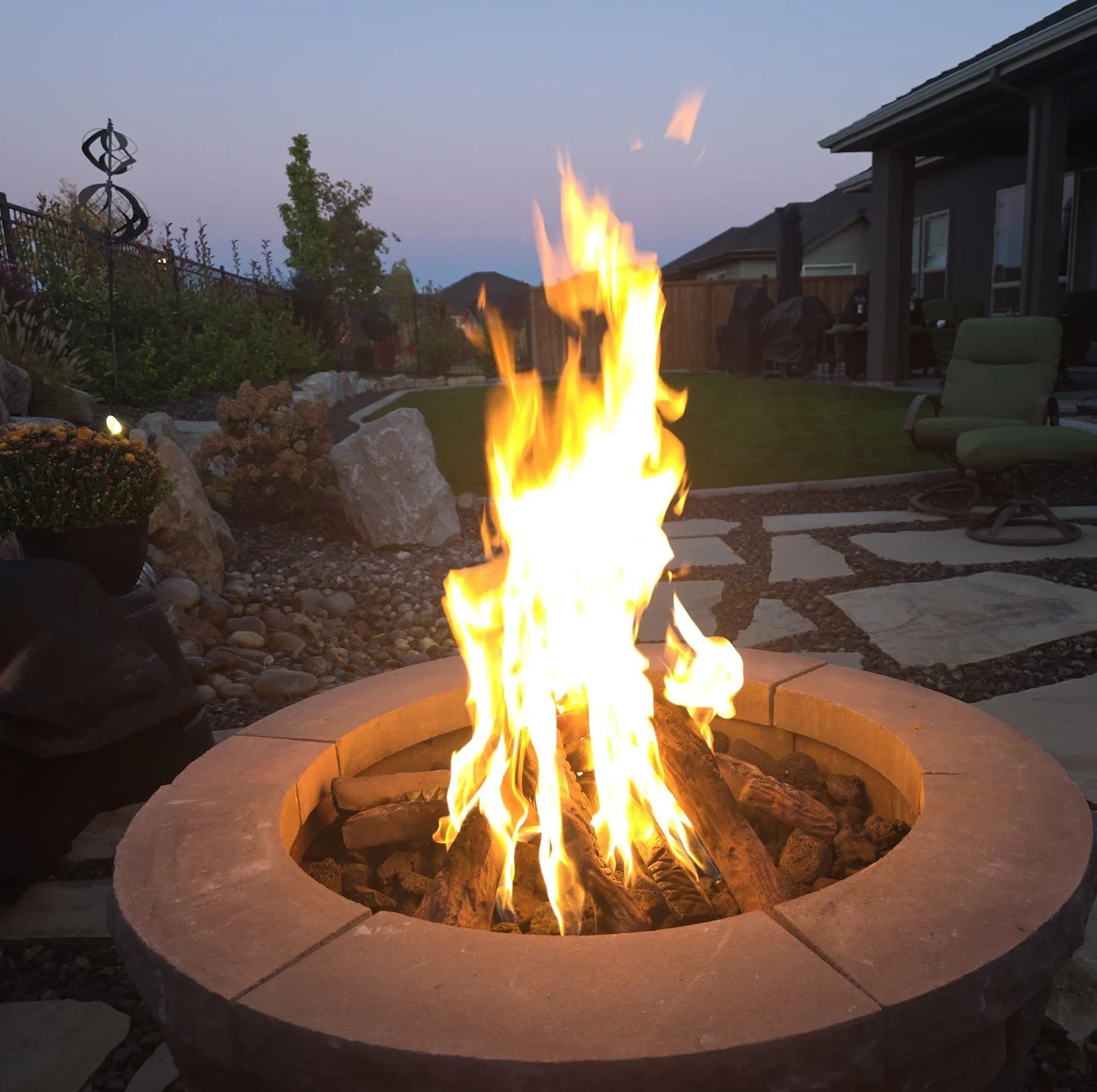 A backyard with a fire pit, lit and burning with bright flames, surrounded by pavers and landscaping, with patio furniture and a house in the background at dusk.