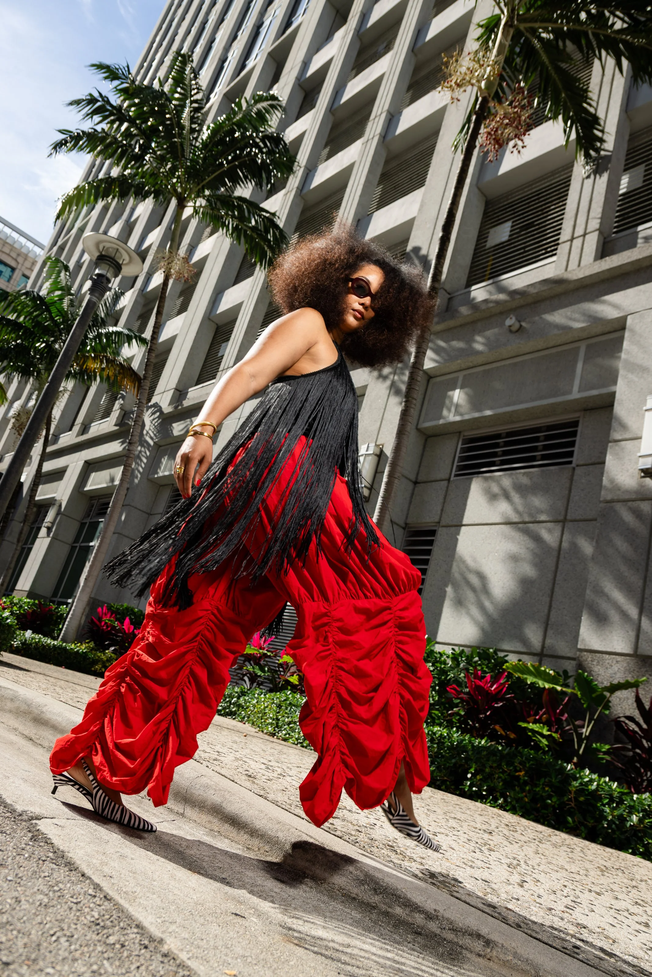 A woman with curly hair and wearing sunglasses walking on a city sidewalk with modern buildings and palm trees in the background.