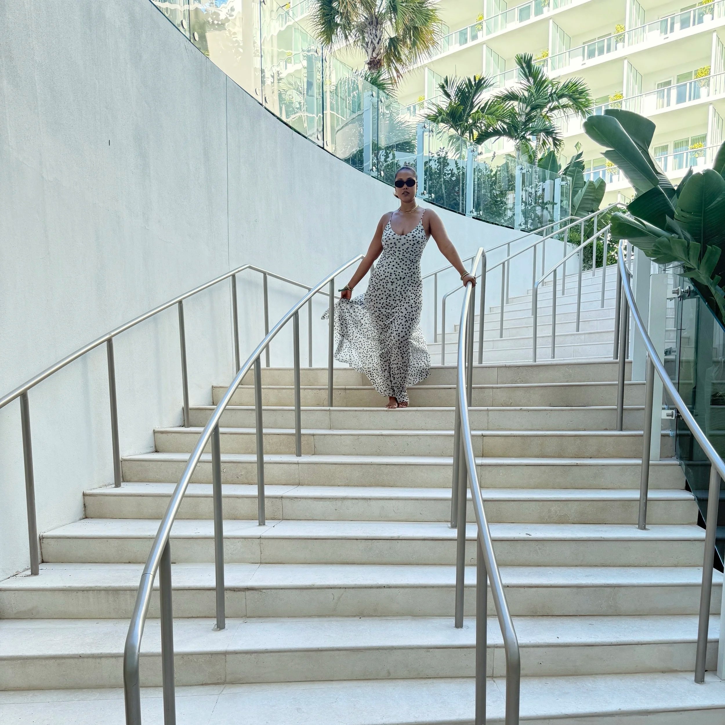 Woman in a polka dot dress walking down outdoor stairs with metal railings, glass building and palm trees in background.