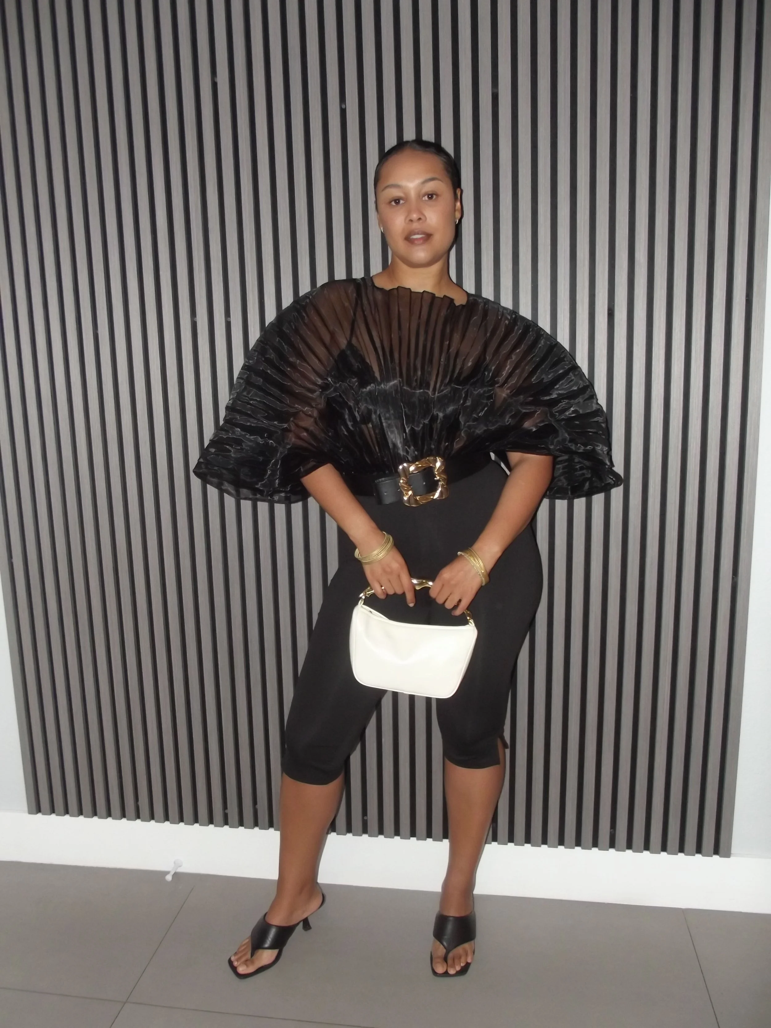 Woman in black outfit with gold jewelry holding a white handbag standing in front of a black vertical striped wall.