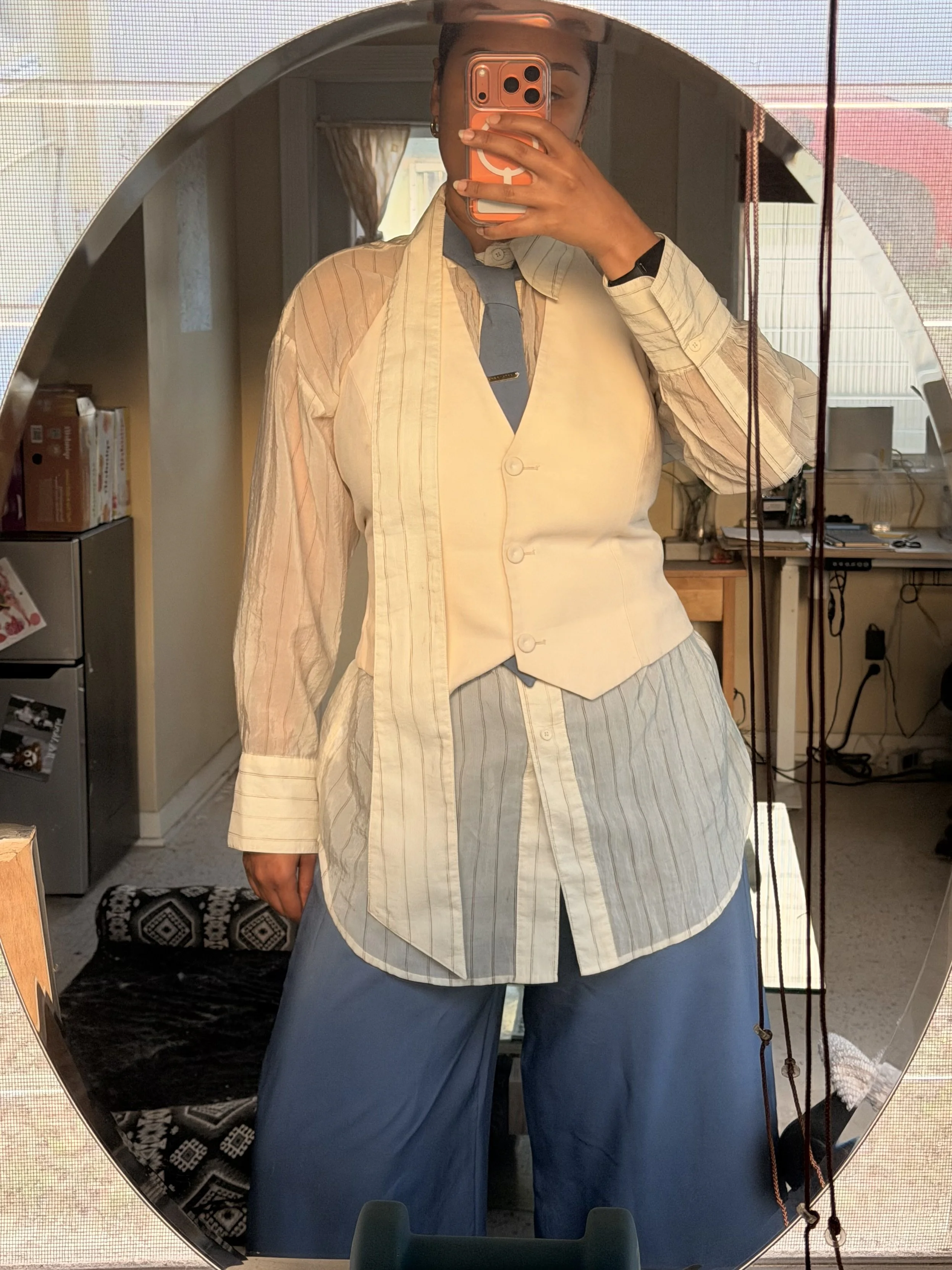 Person taking a mirror selfie, dressed in a beige vest, striped shirt, blue tie, and blue wide-leg pants, holding a pink phone.