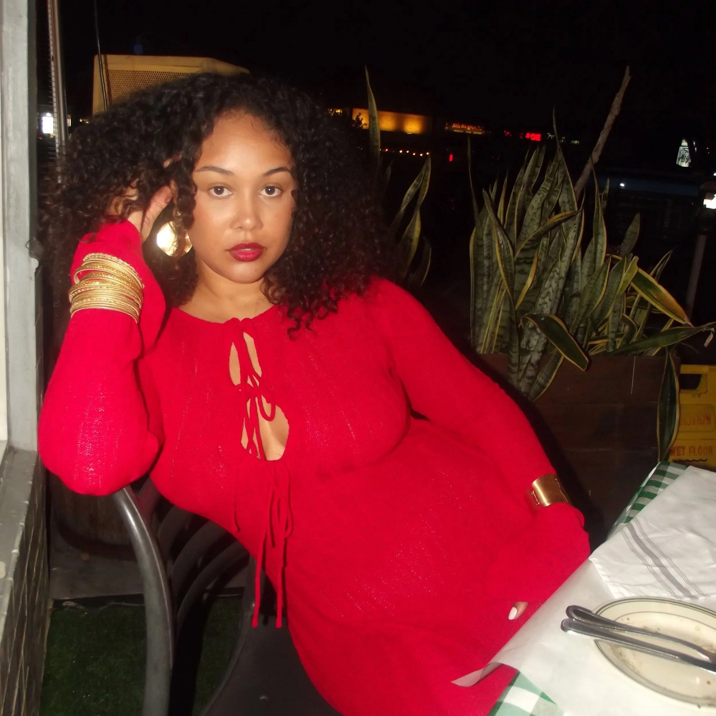 A woman with curly hair wearing a red dress, sitting at a table at night, looking at the camera, with a potted plant in the background.