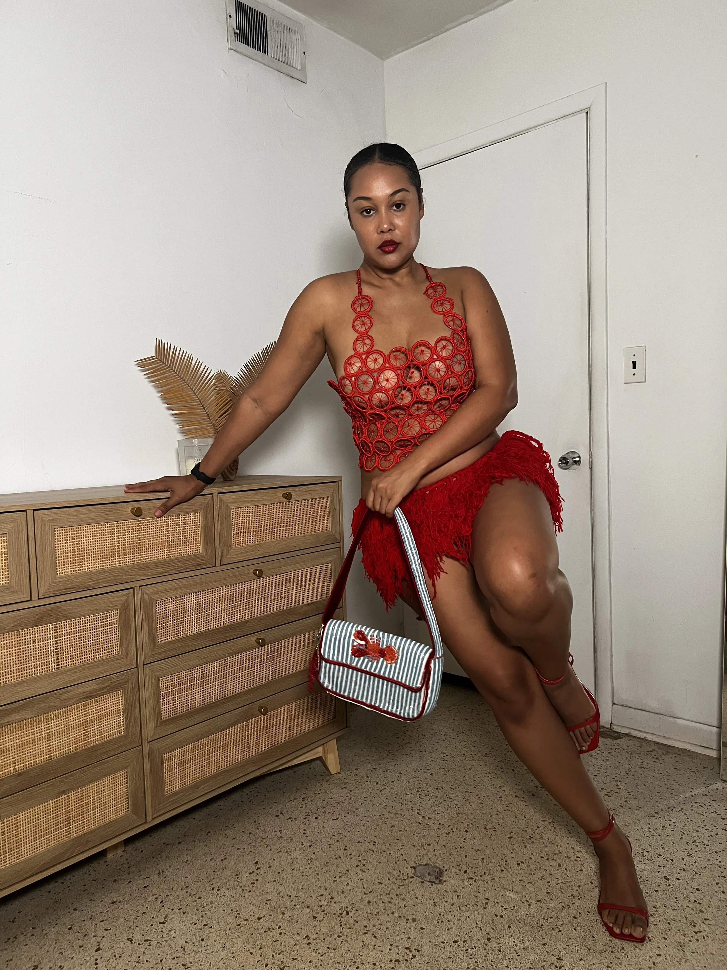 A woman with dark hair tied back, wearing red clothes and red heels, posing indoors with one leg raised and resting her hand on a wooden dresser, holding a striped handbag with a lobster embellishment.