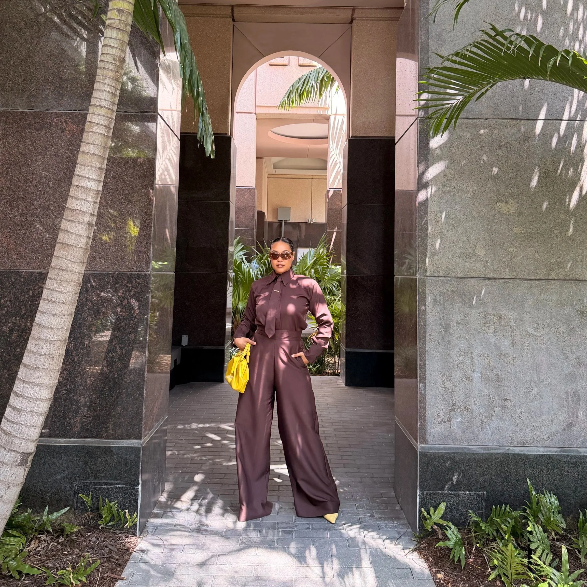 A woman in a shiny brown jumpsuit and sunglasses stands in an arched walkway with plants and modern building architecture around her.