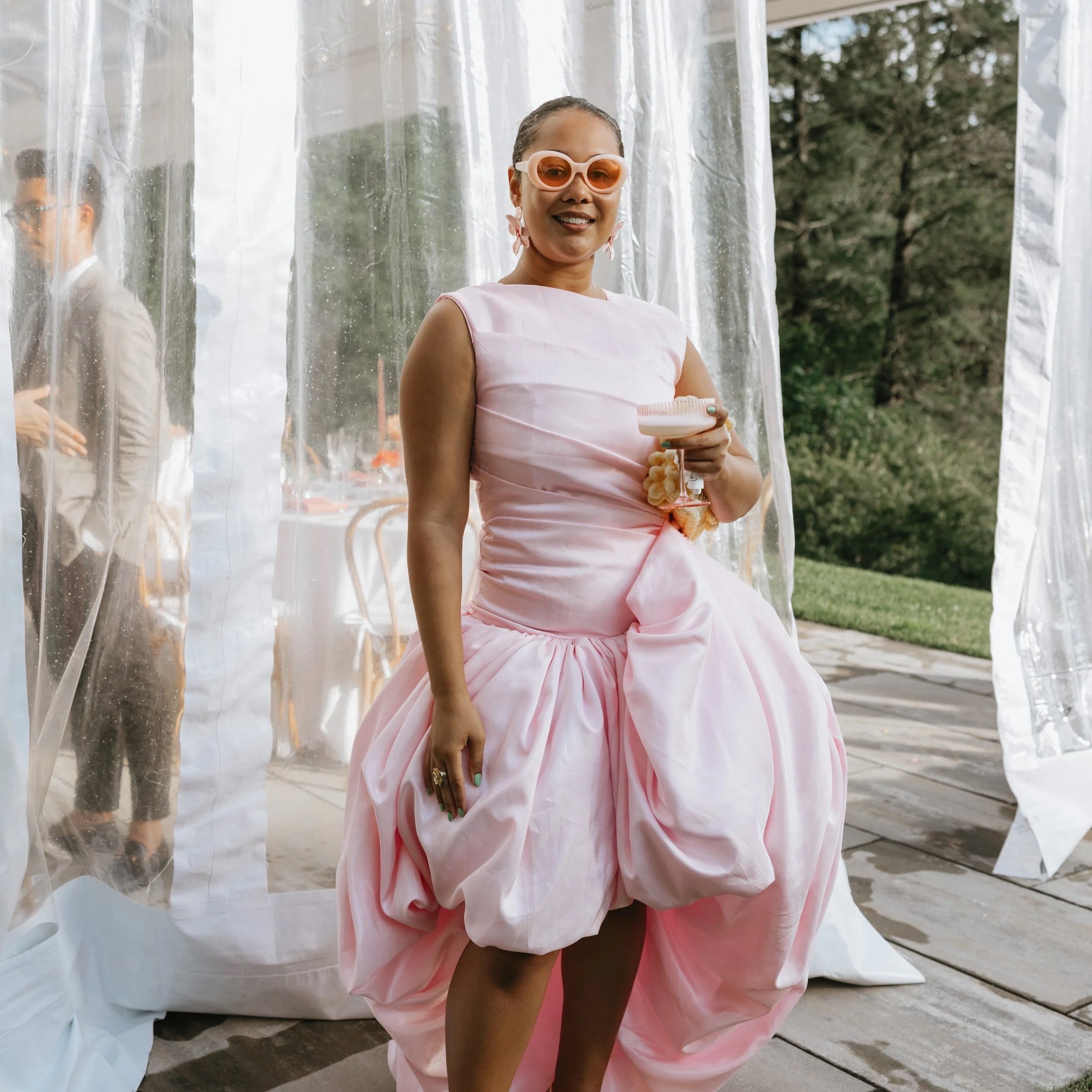 A woman in a pink, puffy dress holding a glass and wearing sunglasses, standing outdoors near a clear plastic curtain.