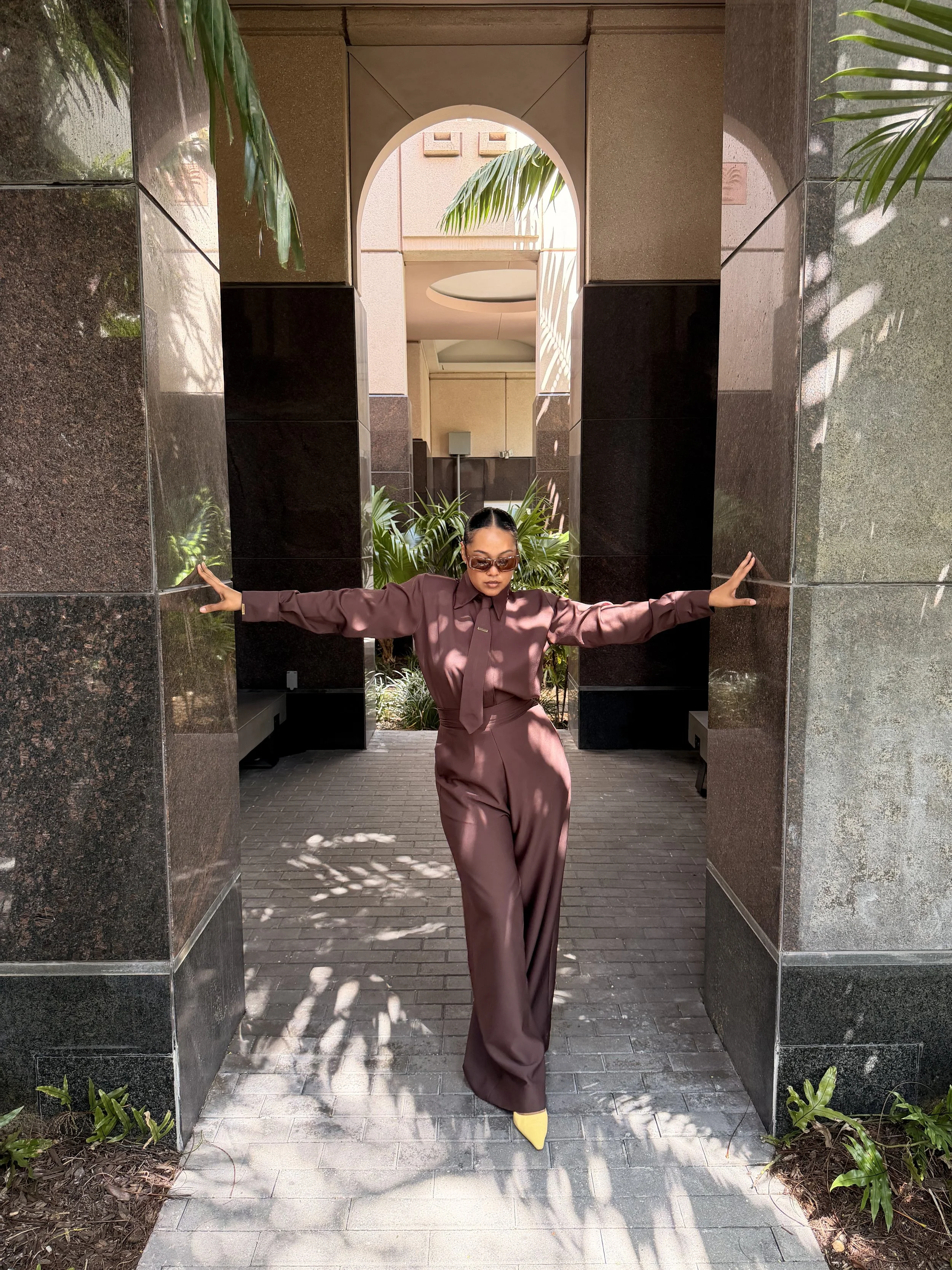 A woman wearing a brown outfit and yellow shoes standing with her arms outstretched in a modern, outdoor architectural setting with plants and shadows.