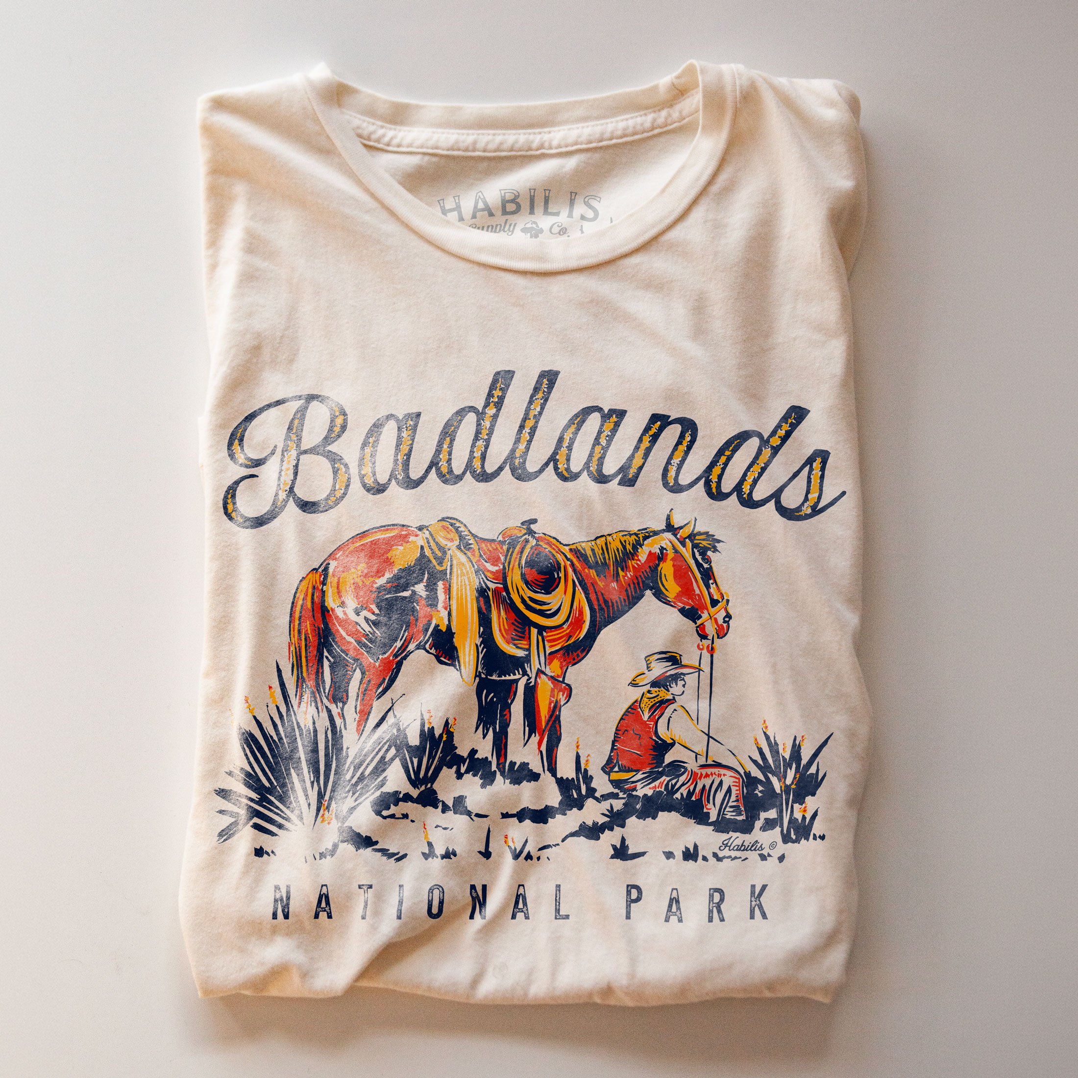 Folded cream T-shirt featuring original Badlands National Park artwork. Photographed on a warm cream background with the graphic prominently displayed.