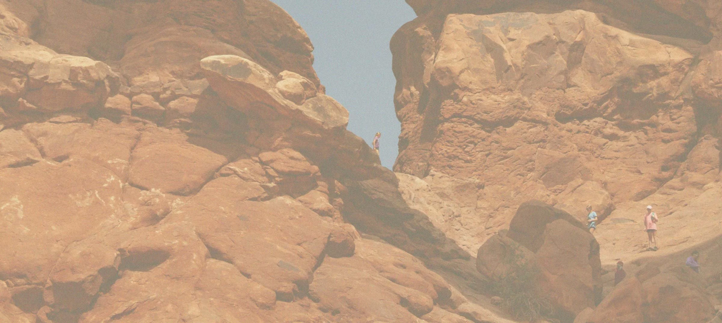People hiking and exploring on a rocky desert canyon with large, reddish-brown rock formations under a clear blue sky.