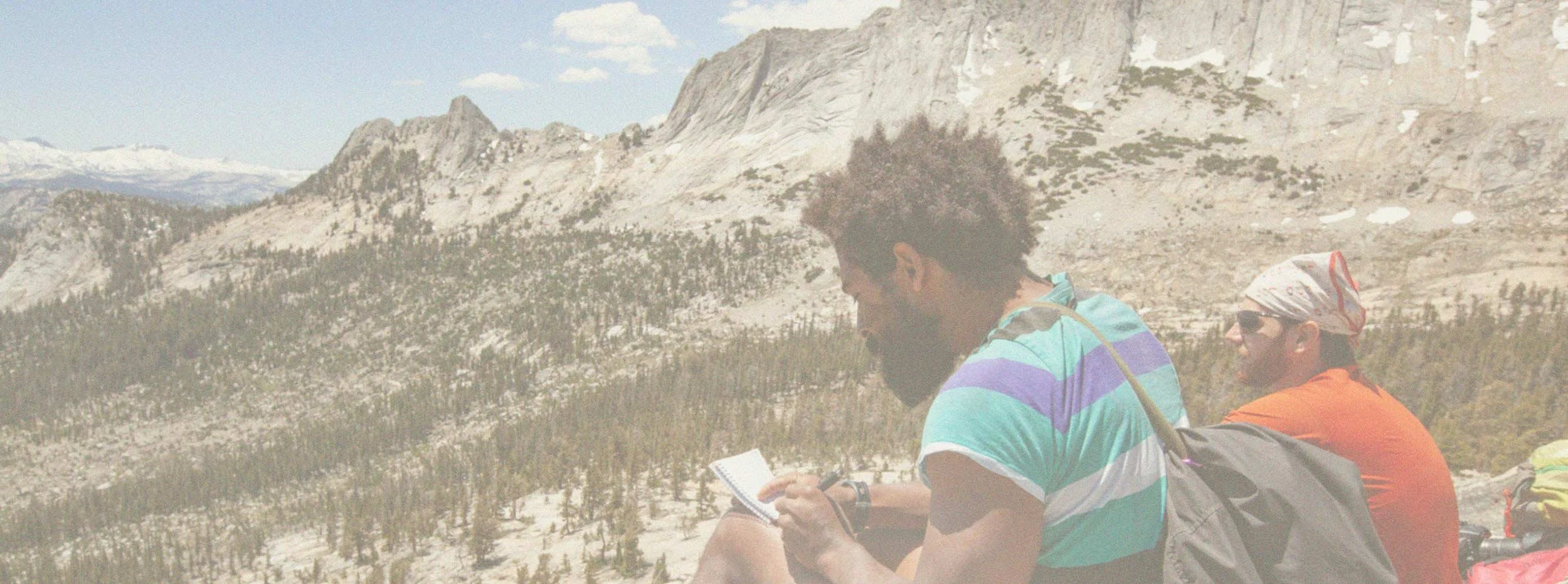 Two men sitting outdoors in a mountainous landscape, one reading a book and wearing a striped shirt, the other in a red shirt with sunglasses and a bandana, with backpacks nearby.