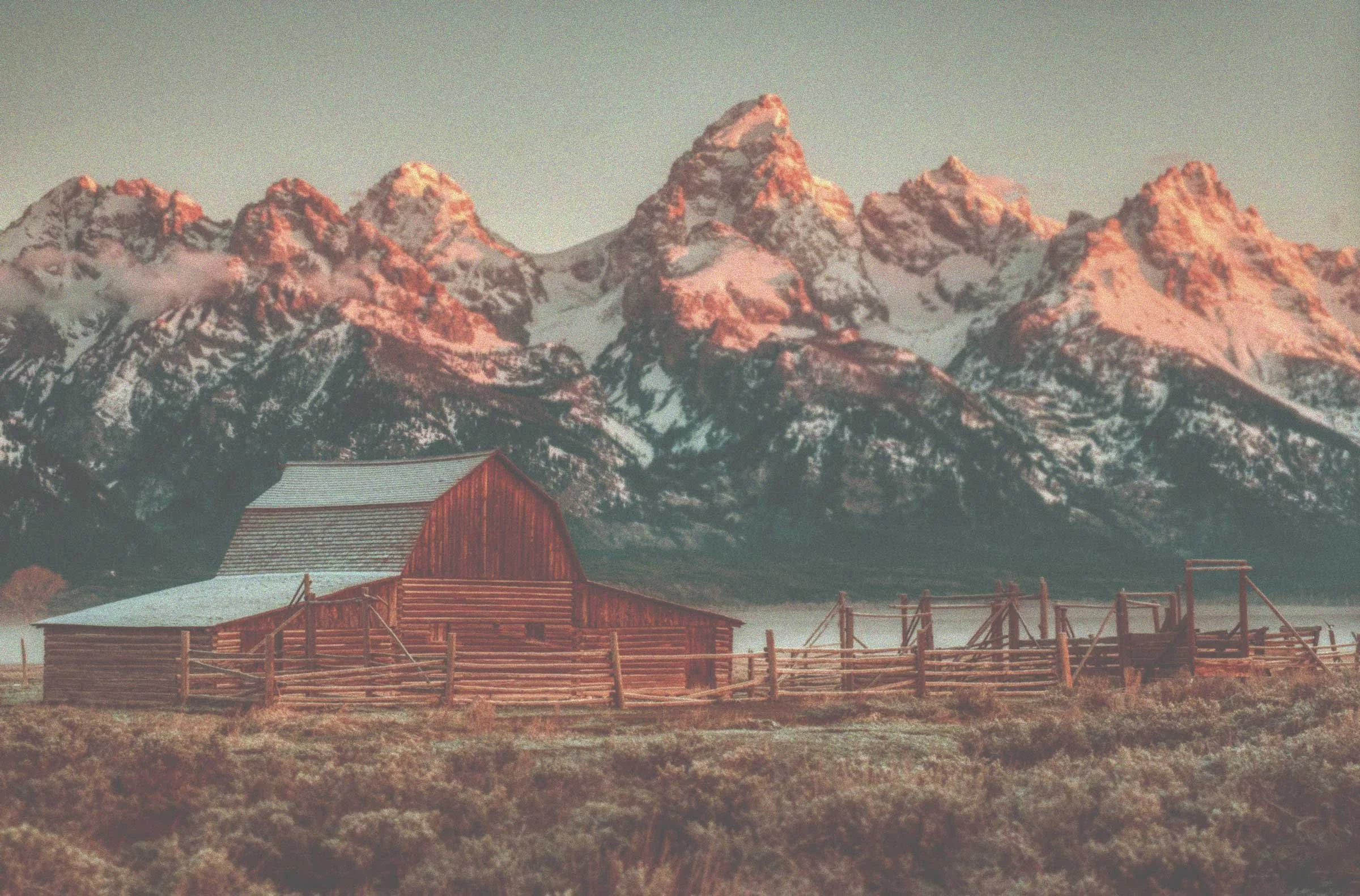 A rustic wooden barn in a field with mountains in the background, during sunrise or sunset.