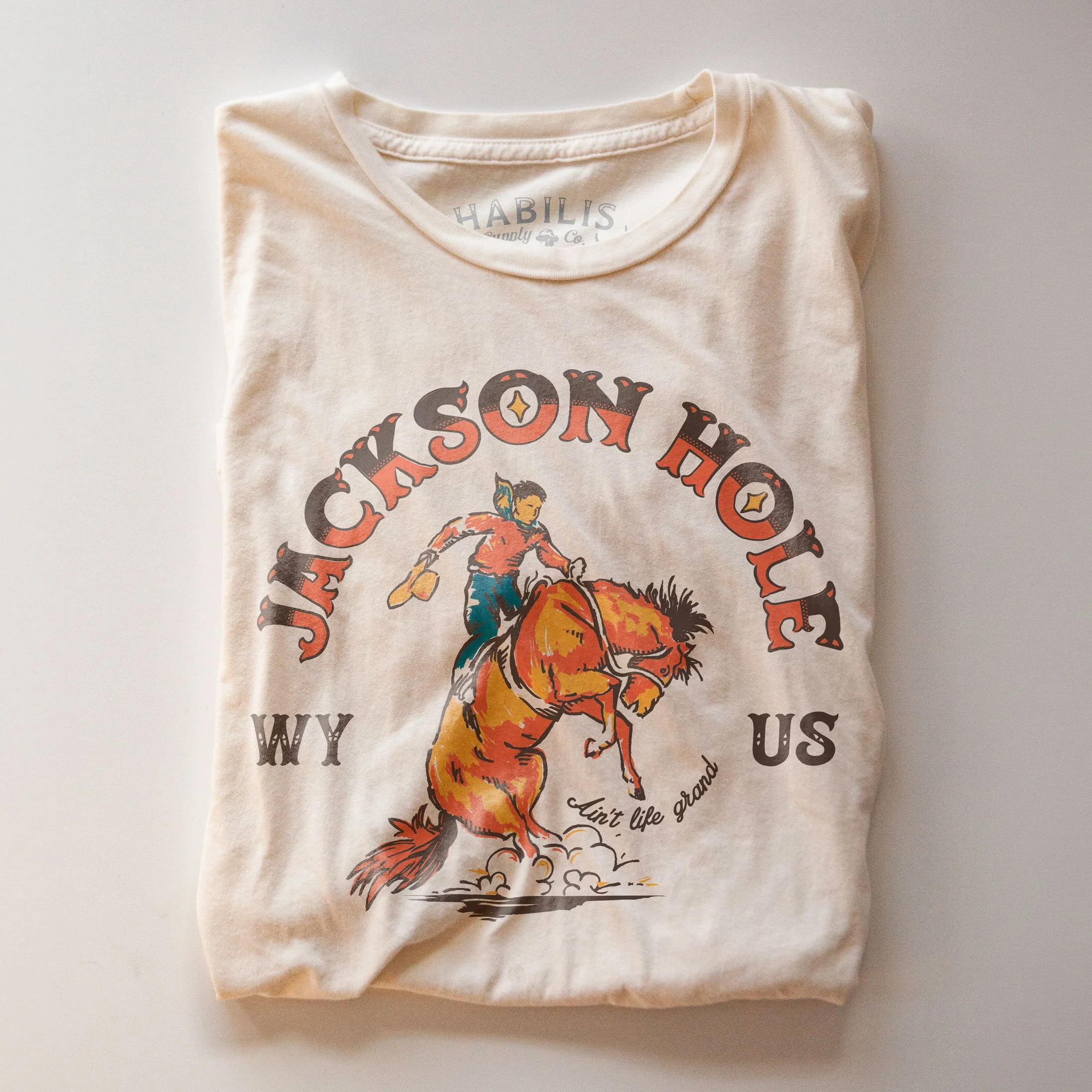 Folded cream T-shirt featuring original Jackson Hole artwork. Photographed on a warm cream background with the graphic prominently displayed.