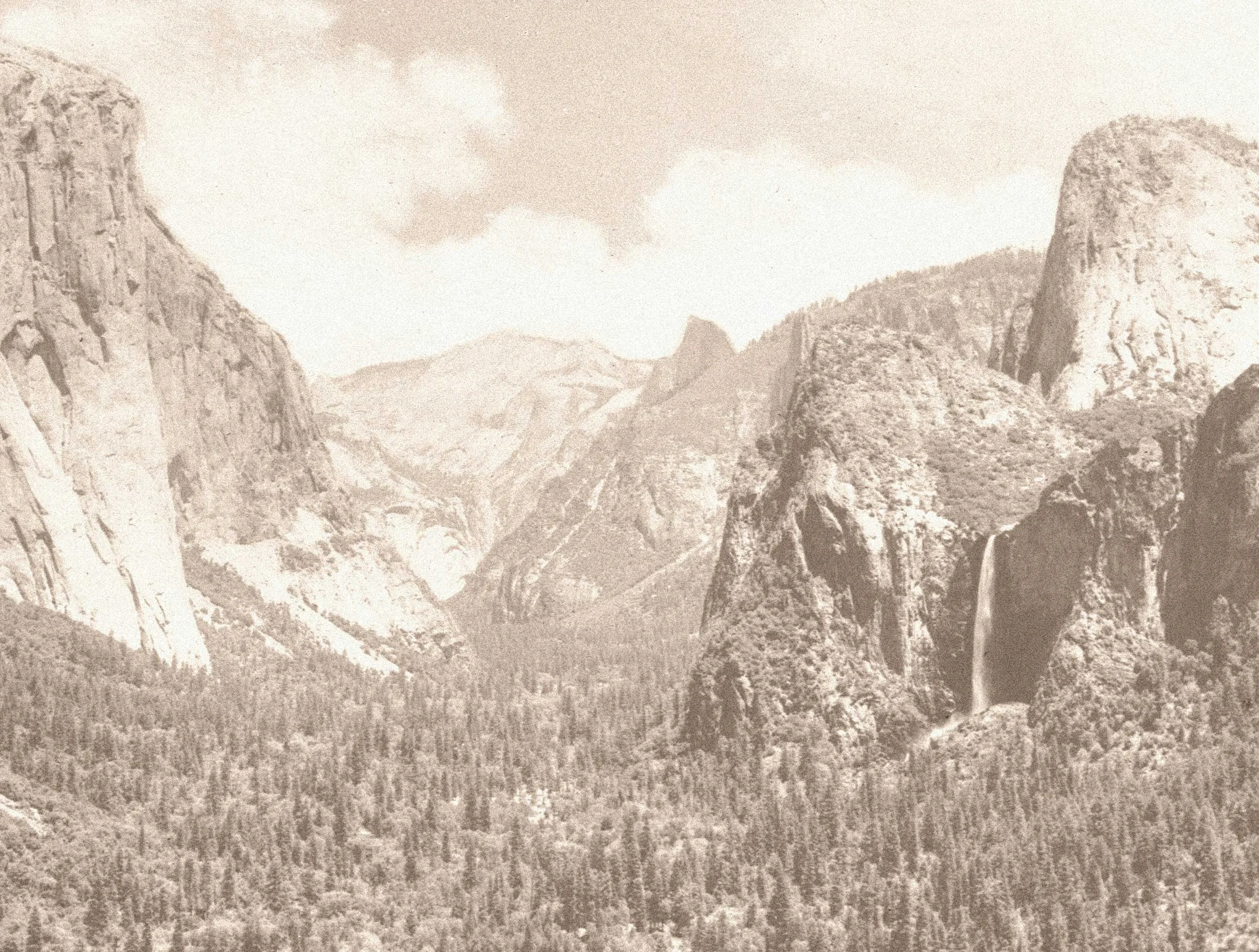Sepia-toned photograph of a mountainous landscape with tall peaks, dense forest in the foreground, and a waterfall cascading down a cliff on the right.