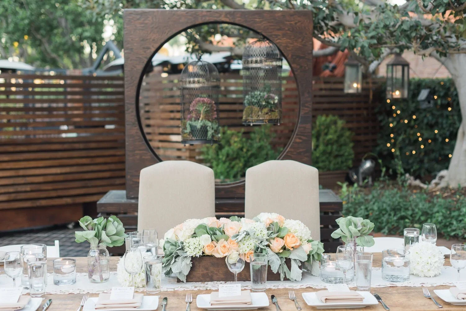 Outdoor wedding reception setup with a decorated dining table, floral centerpieces, and hanging lanterns with candles, surrounded by greenery and a wooden fence.