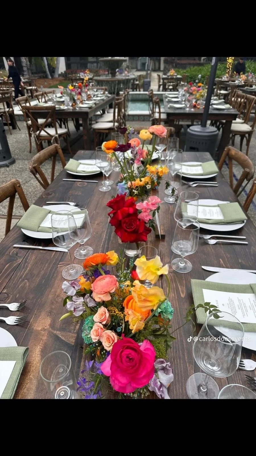 A long wooden table set for a meal with colorful flower arrangements as the centerpiece, green napkins, white plates, silverware, and empty wine glasses, outdoors with additional tables and chairs in the background.
