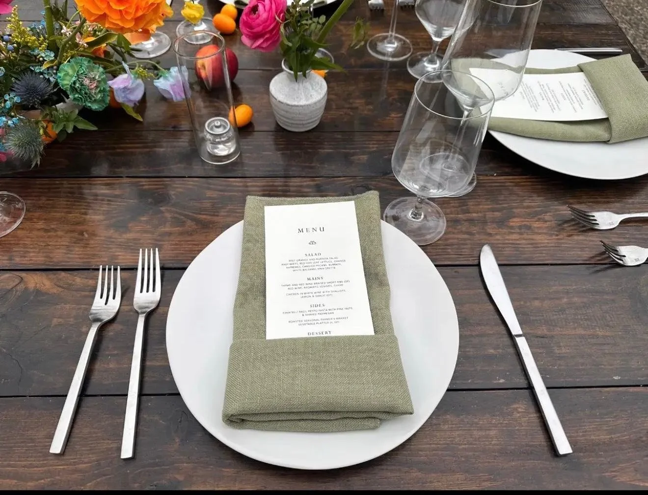Dining table setting with white plate, green cloth napkin, printed menu, three forks, knife, empty wine glasses, and floral centerpiece on a dark wooden table.