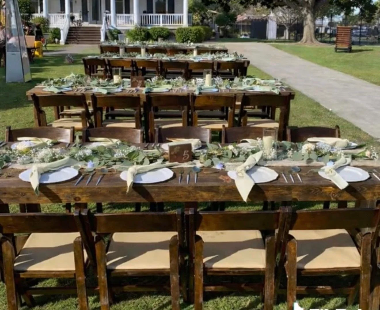 Outdoor dining setup with long wooden tables decorated with white plates, napkins, silverware, floral centerpieces, and candles in a grassy area.