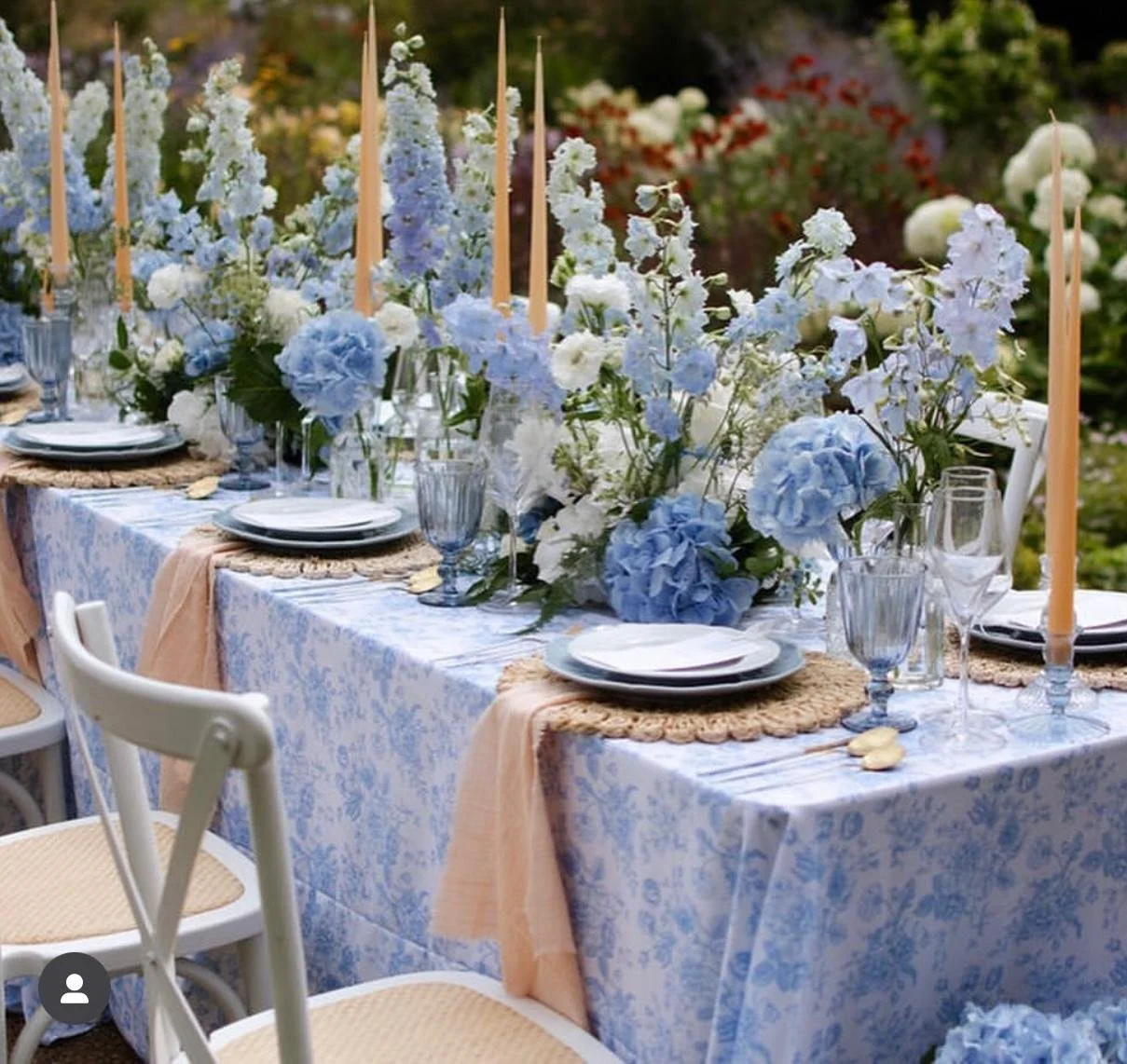 Elegant outdoor table setting with blue and white flowers, tableware, and tall candles.