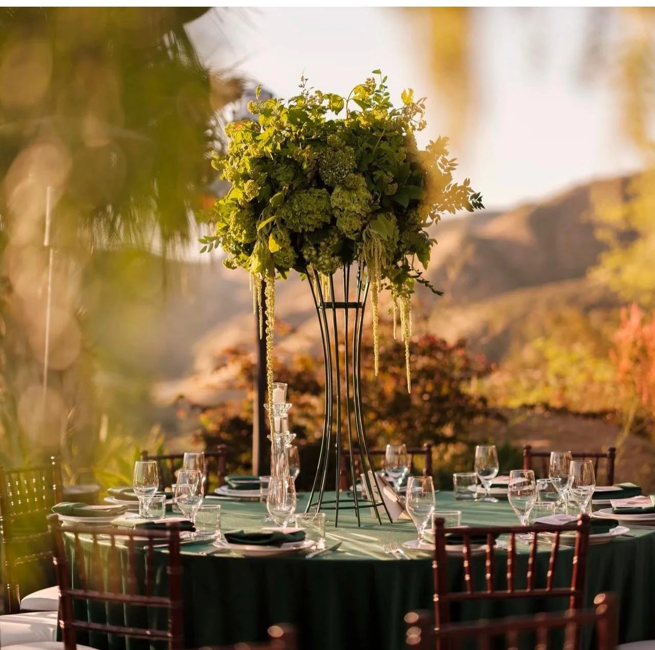 Elegant outdoor dining table set with wine glasses, plates, and cutlery, featuring a tall floral centerpiece on a green tablecloth, with a sunset and mountain landscape in the background.