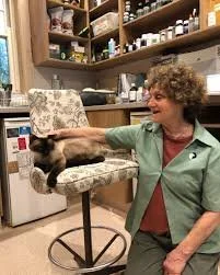 Woman with curly hair smiling at a cat on her lap in a room with shelves and equipment.