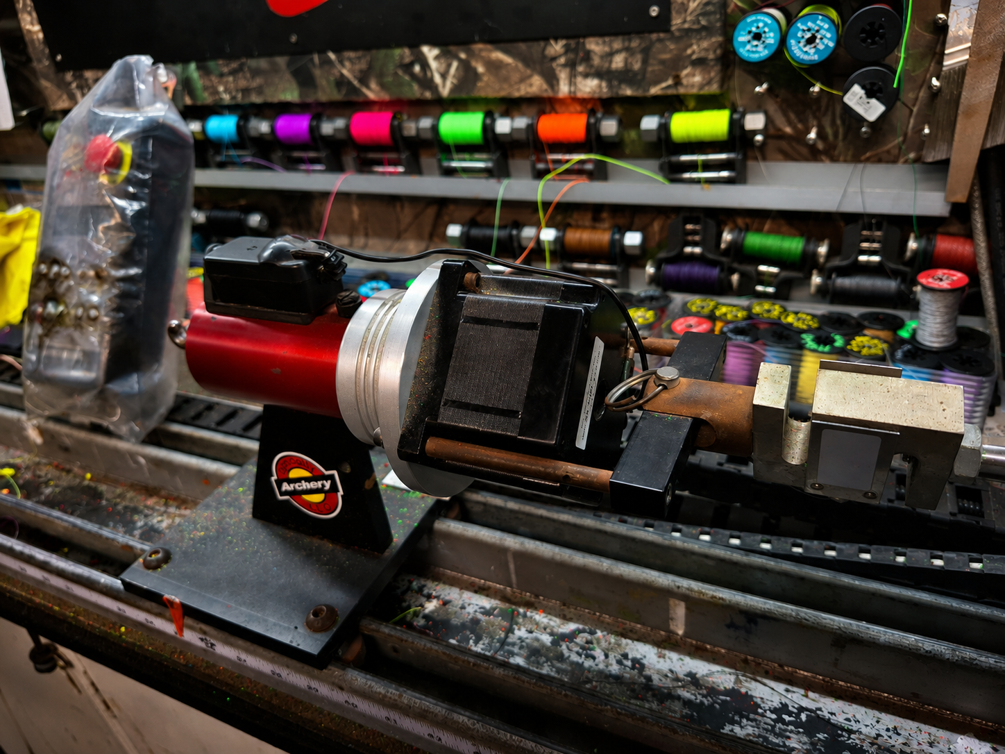 A machine for making archery arrows on a workbench with spools of colorful thread and materials in the background.