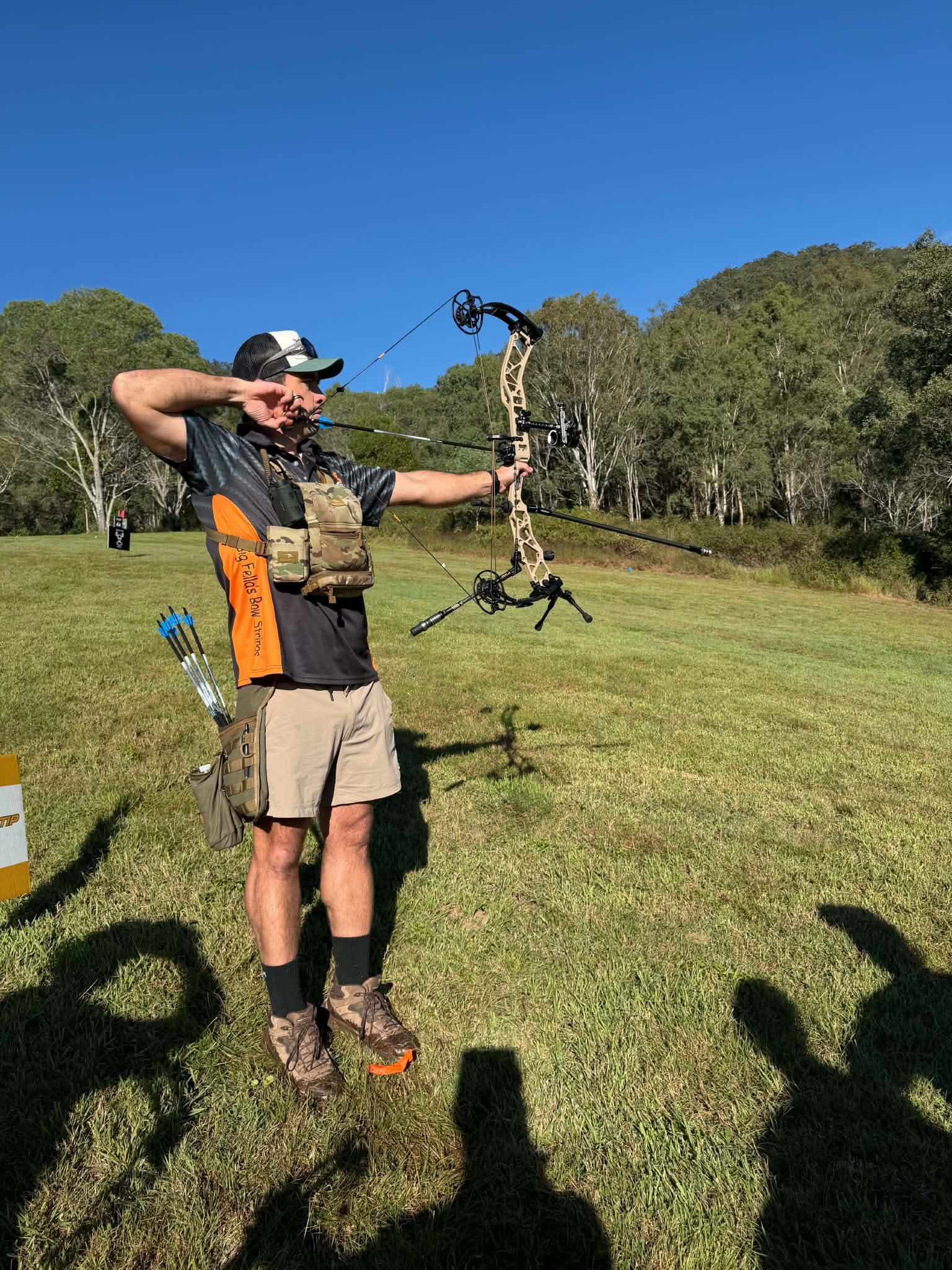 Man practicing archery outdoors on a grassy field, aiming a compound bow with a bright yellow arrow, with trees and a clear blue sky in the background.
