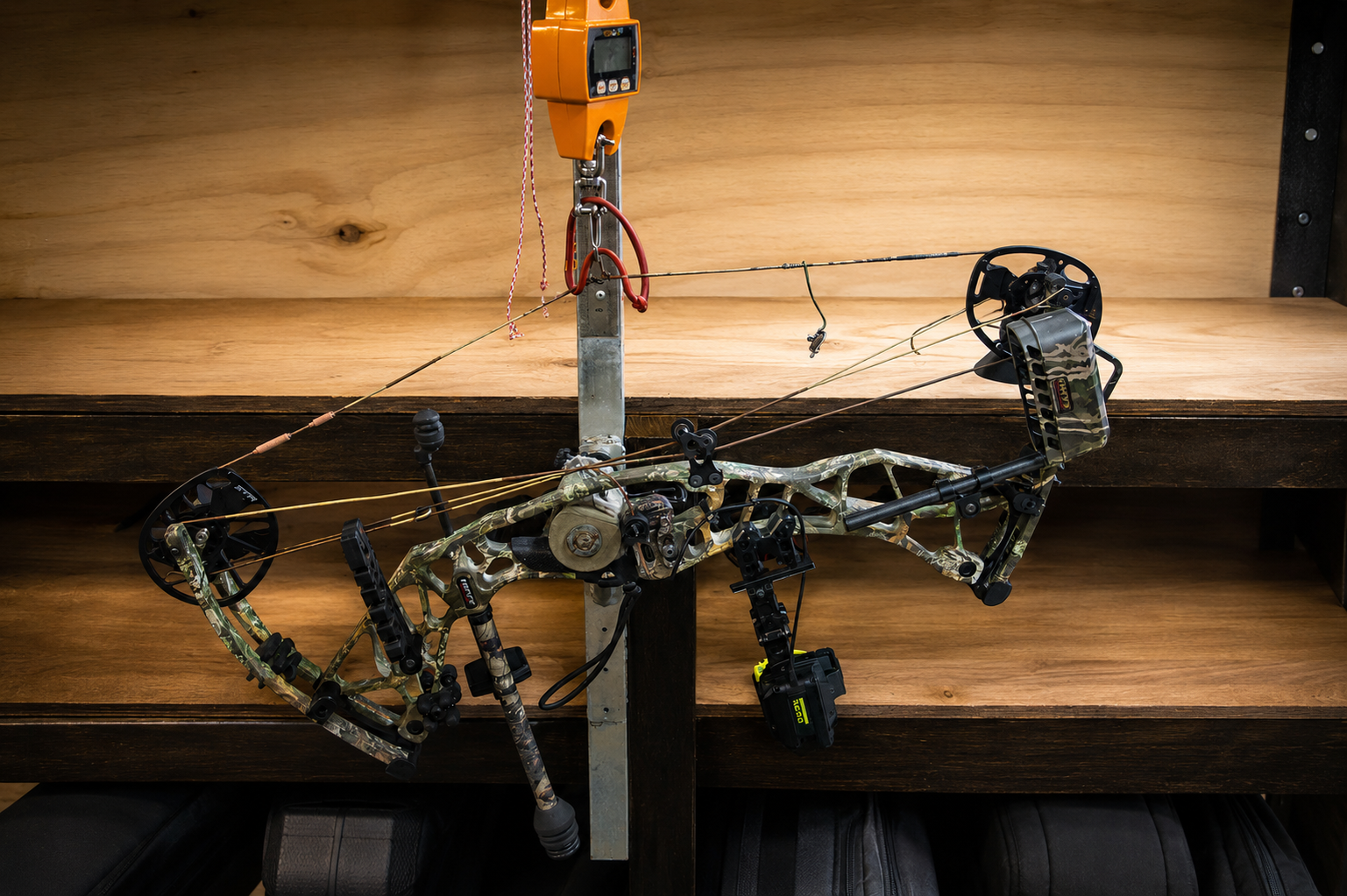 A camouflage-patterned compound bow mounted on a wooden wall with a rope / scale and a black digital device attached.