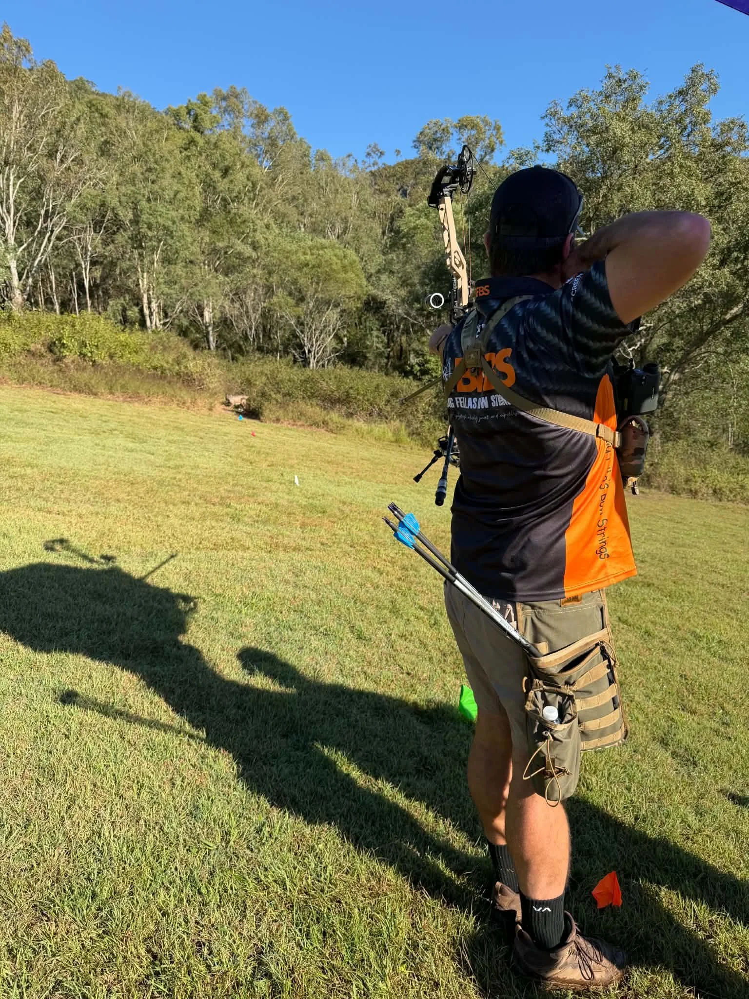 A person aiming a bow and arrow in an outdoor grassy field with trees and a clear blue sky in the background.