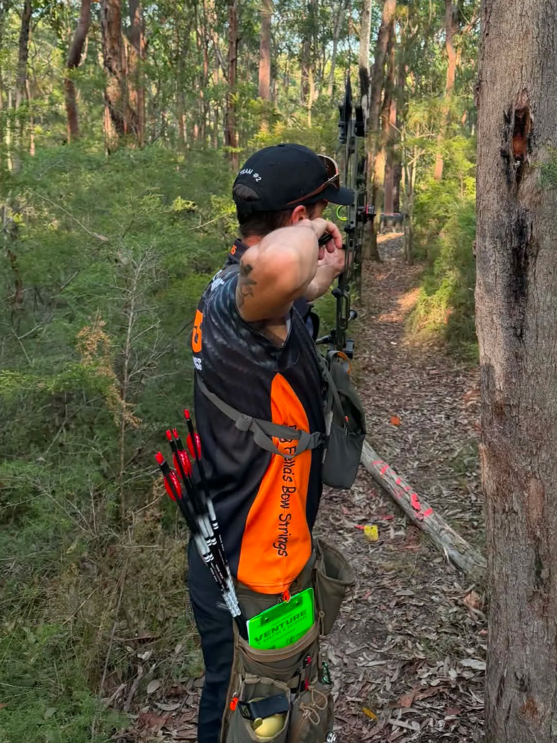 A man practicing archery in a wooded area, drawing a bow and aiming at a target down the trail.