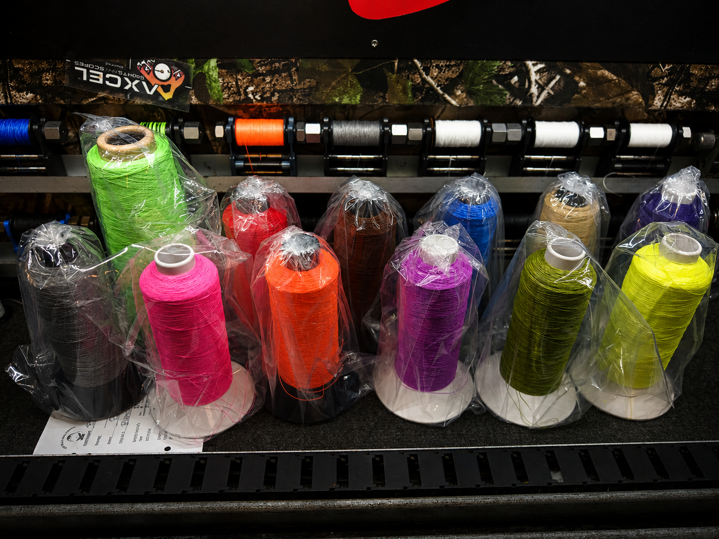 Spools of colorful thread in plastic wrapping on a black surface, with spools of more thread and ribbons in the background.