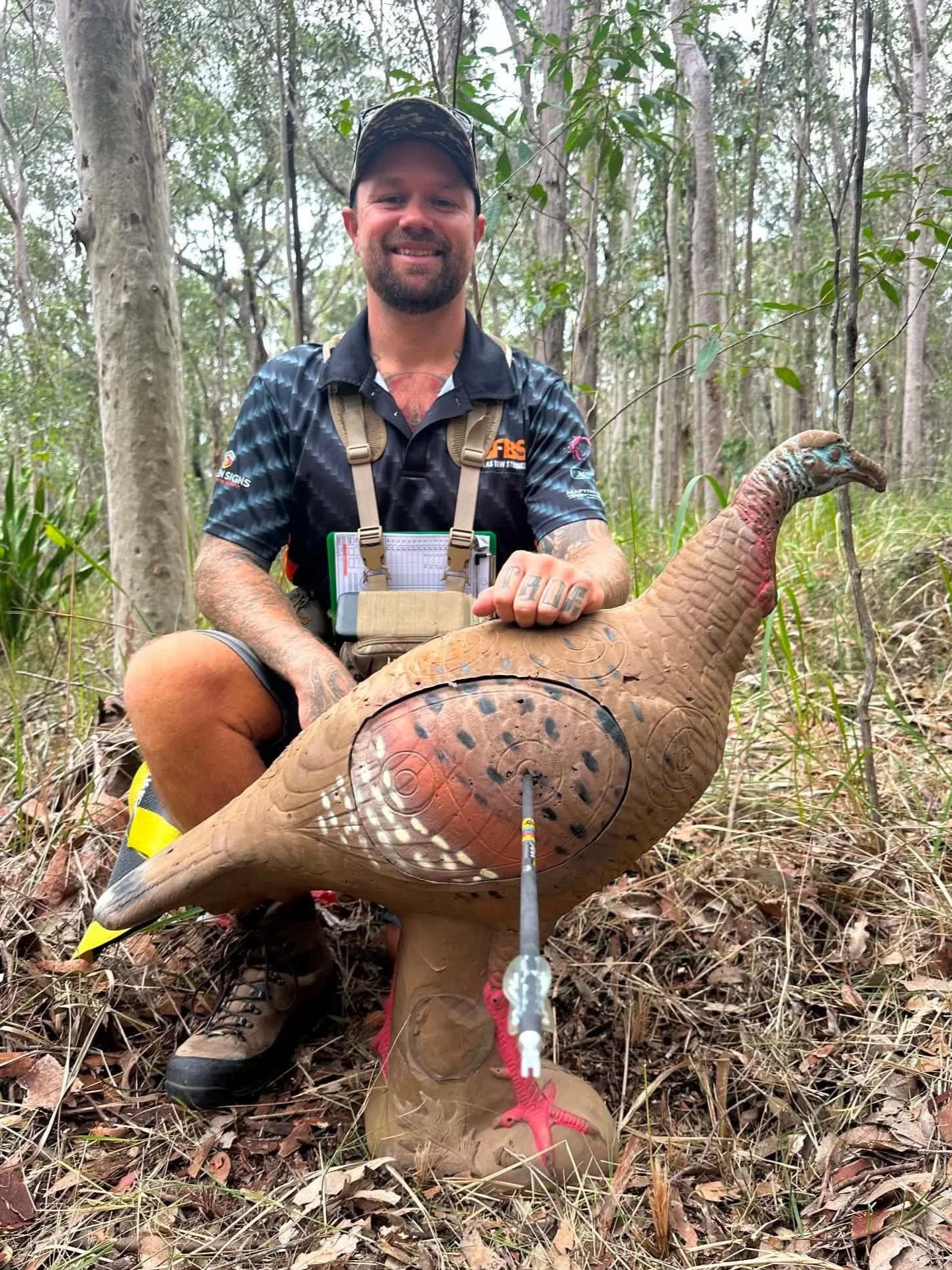 A man with tattoos wearing outdoor gear and a camouflage hat sitting in a forest next to a large, realistic-looking carved or artificial dinosaur figure with a dart in its body.