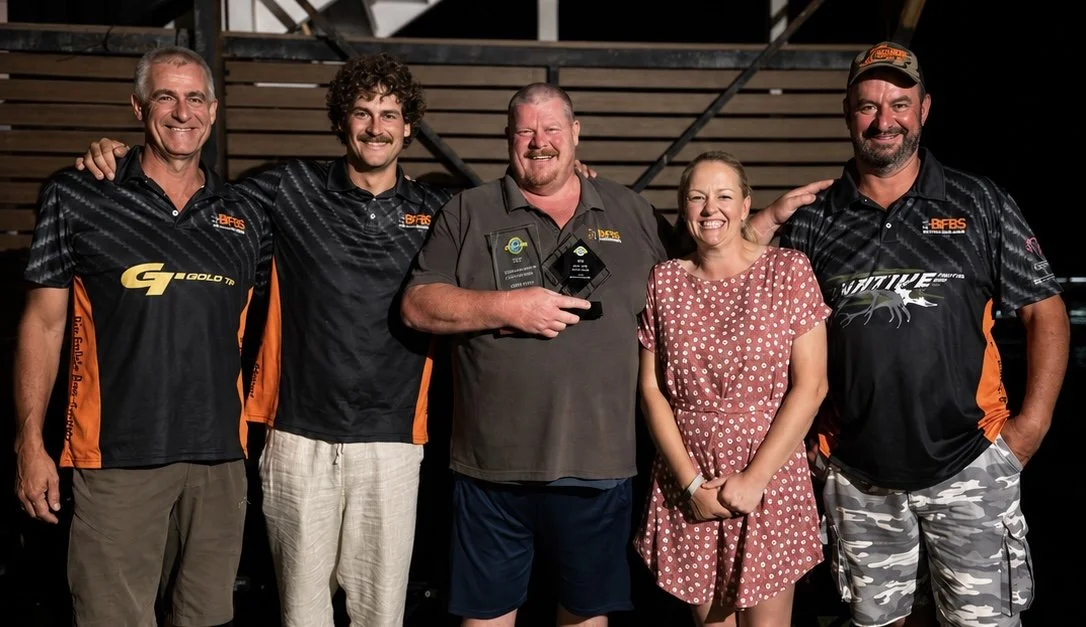 Group of five people standing together, smiling, with some wearing matching sports jerseys, and one holding awards, in front of a wooden backdrop.