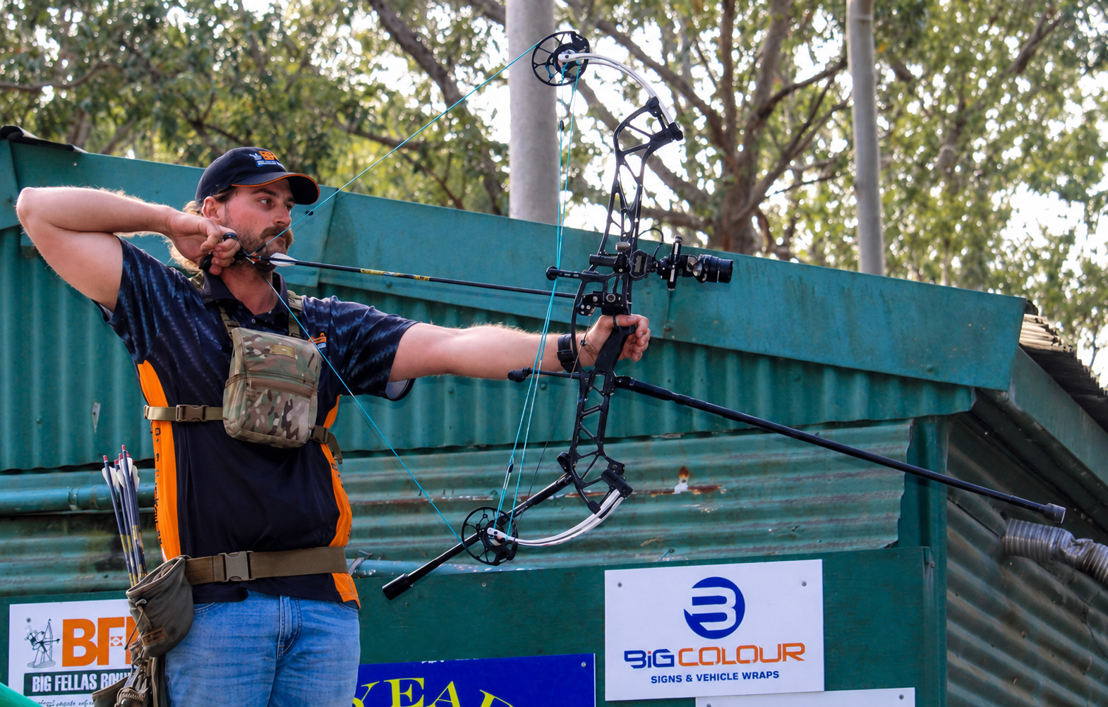 An archer in a BFBS sponsored shirt, shooting a compound bow outdoors. He is aiming at a 3D target.