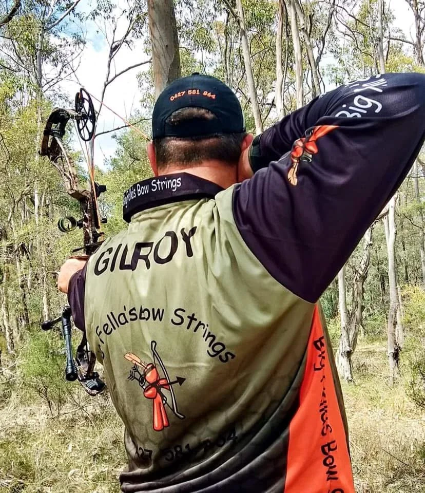 A man outdoors in a wooded area practicing archery, aiming a compound bow. He is wearing a shirt with "GILROY" and "Fella's Bow Strings" written on the back, along with a logo of a bow and arrow.