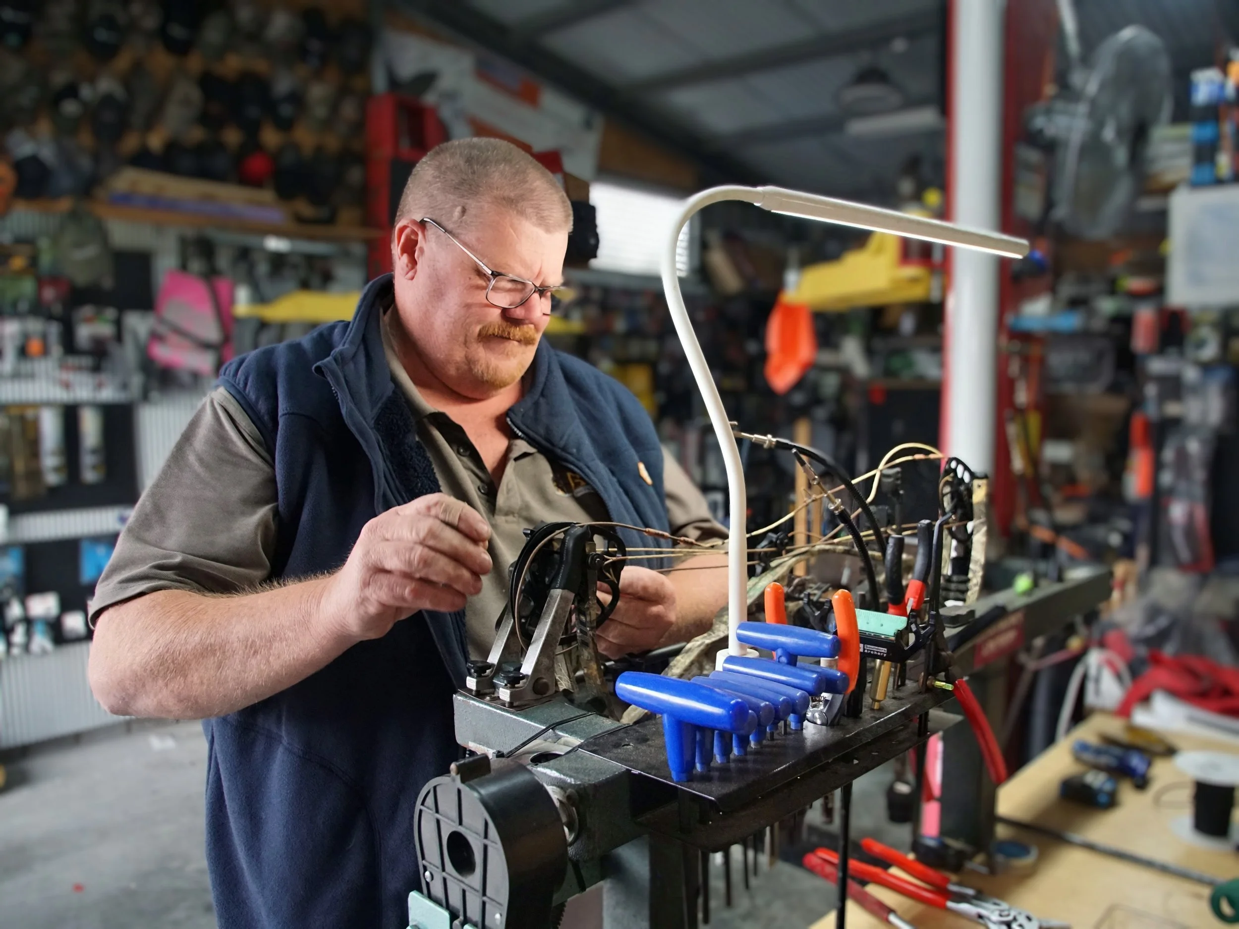 A man with glasses and a short haircut working in a workshop, assembling or repairing a device with wires and tools, surrounded by shelves with various tools and supplies.