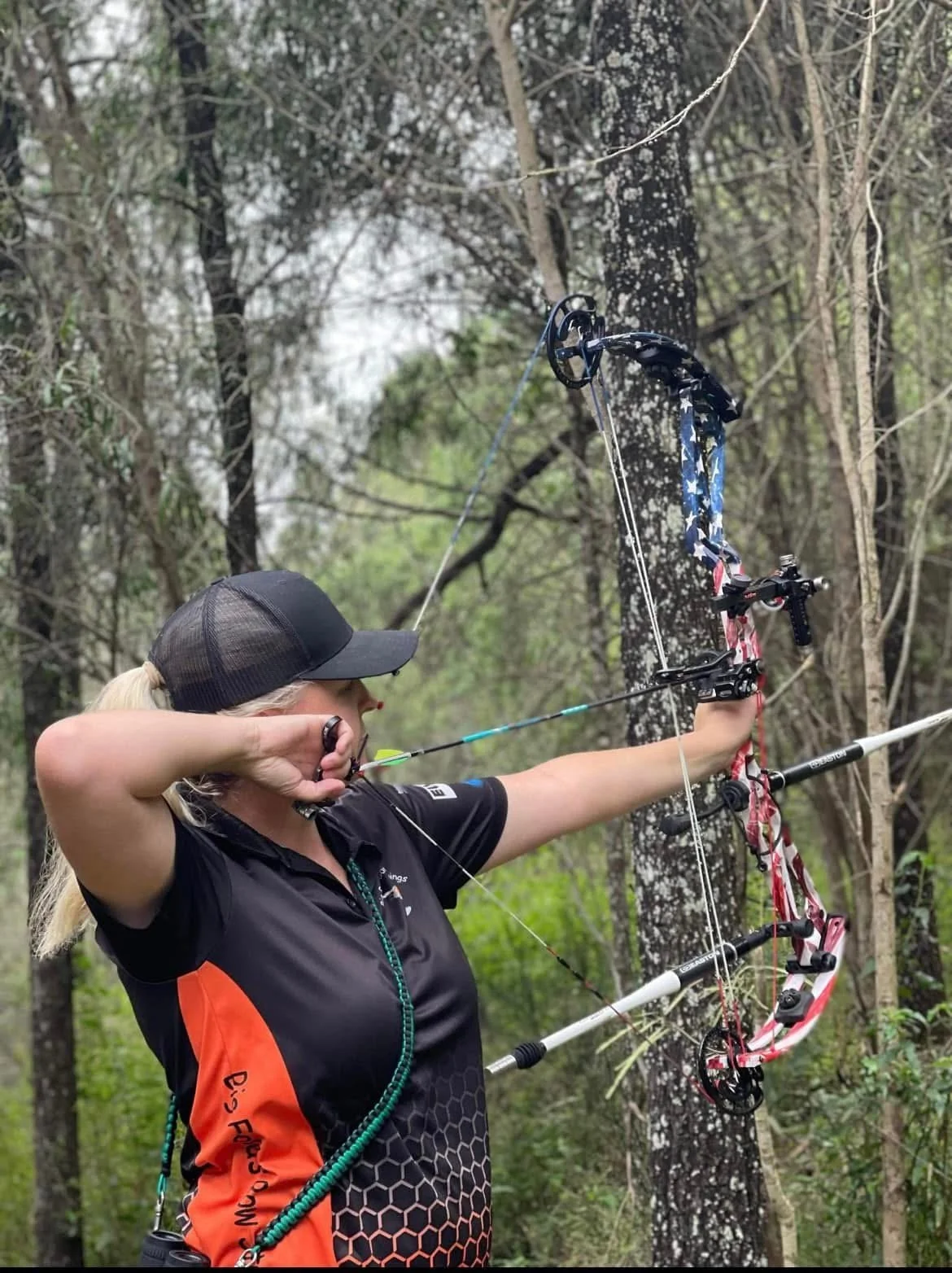 A woman in a black and orange sports shirt and a black cap is aiming a compound bow with a blue and red patterned bowstring in a wooded area. She is pulling back the bowstring with her right hand and holding the bow in her left hand, standing among trees with green leaves.