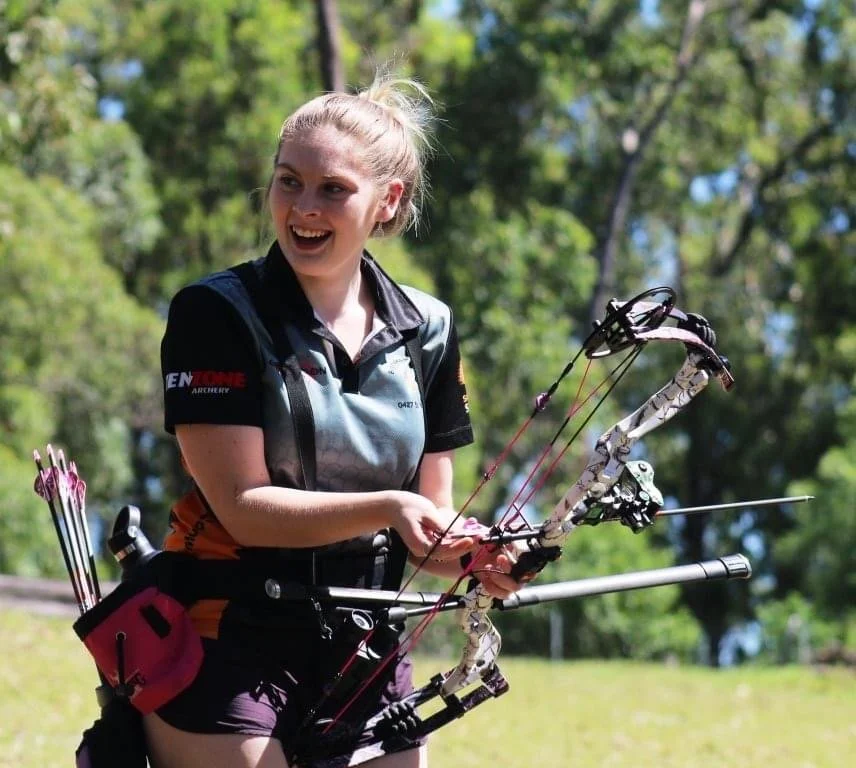 A young woman outdoors, smiling, holding a compound bow and arrows in a wooded area.